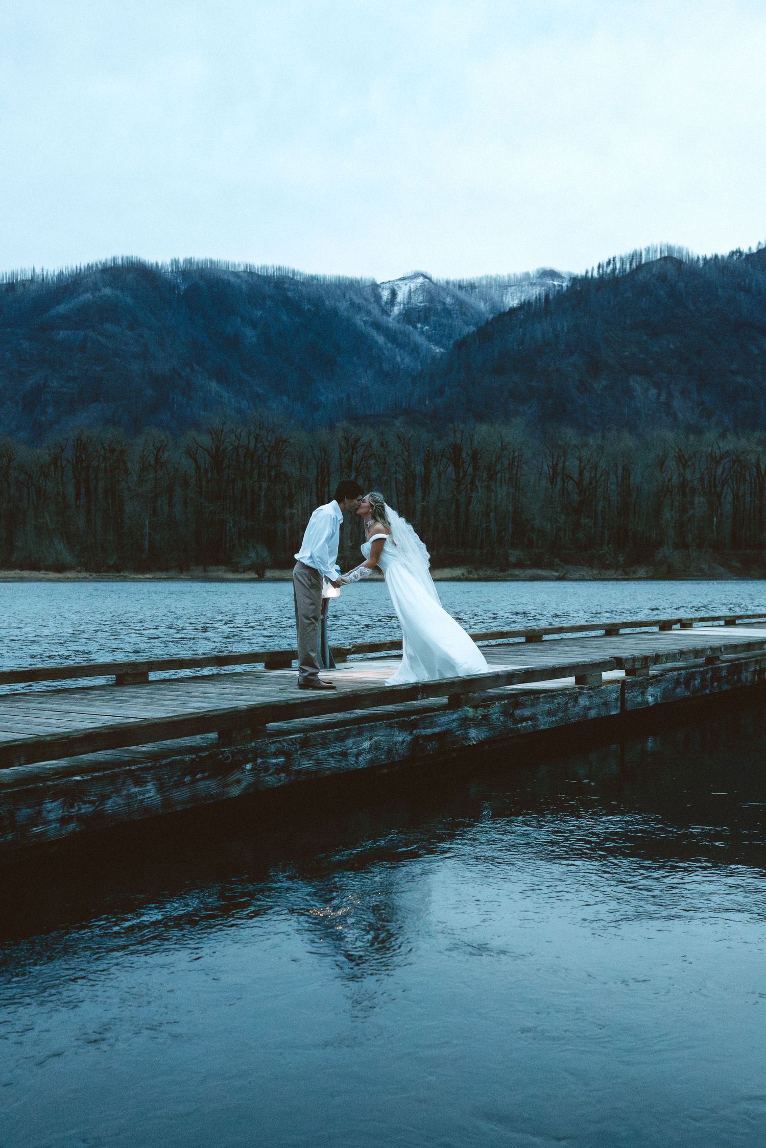Dramatic waterfront wedding photos on a dock with snow-capped mountains in the background.