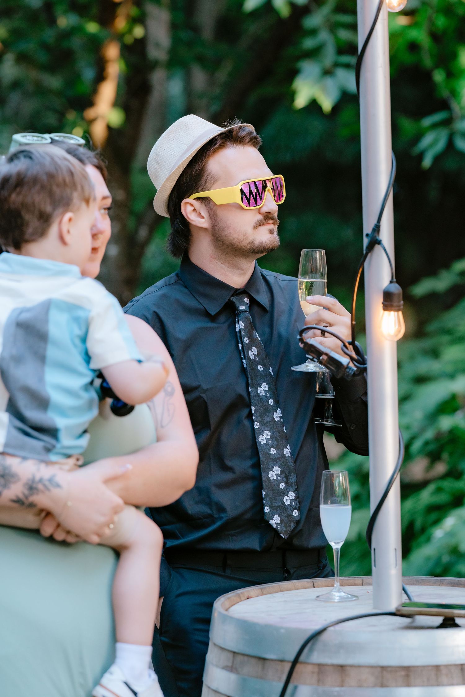 Man in hat and yellow sunglasses holding champagne glass at outdoor wedding reception near barrel.