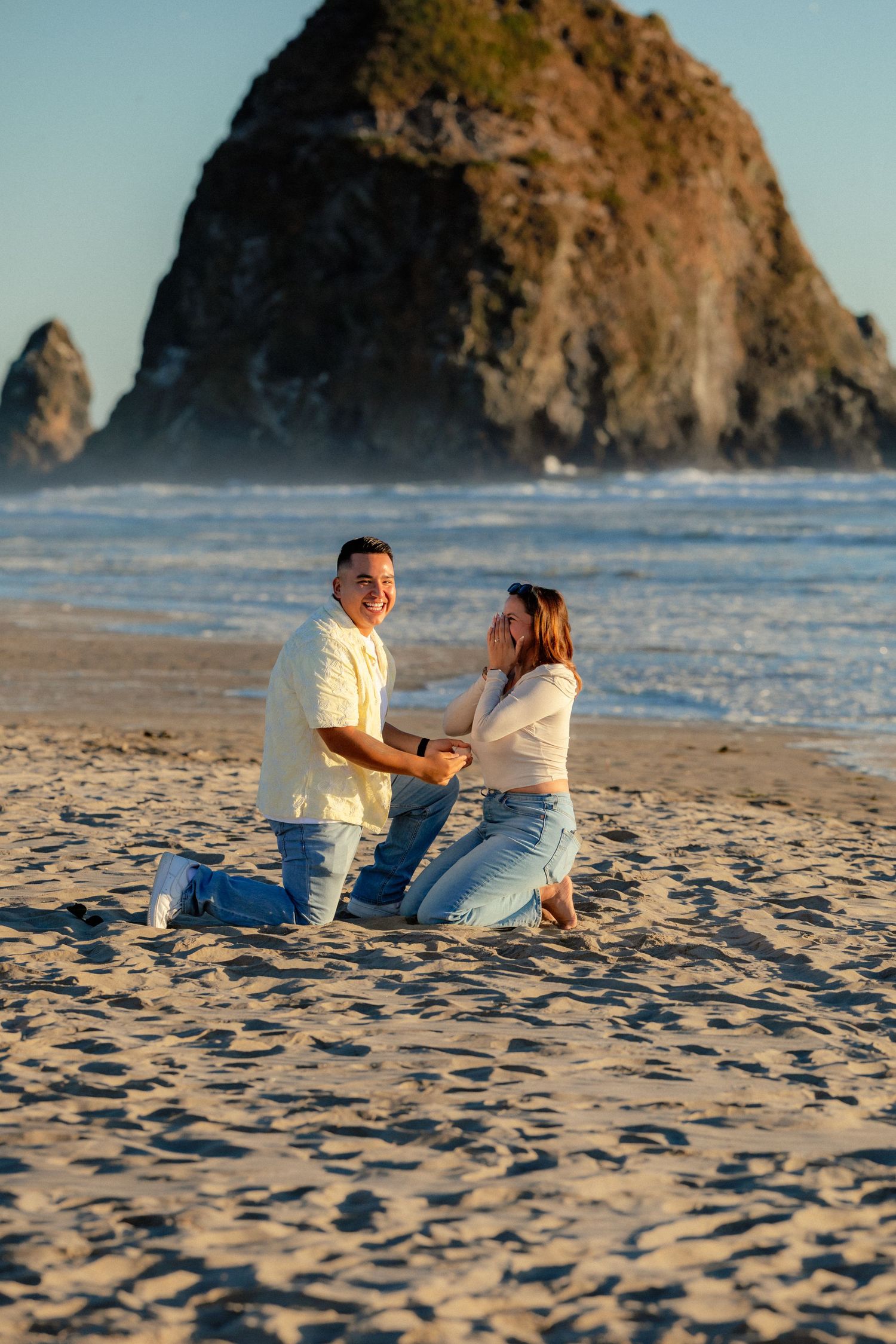 Joyful marriage proposal on Cannon Beach with man on knee holding hands with emotional woman.
