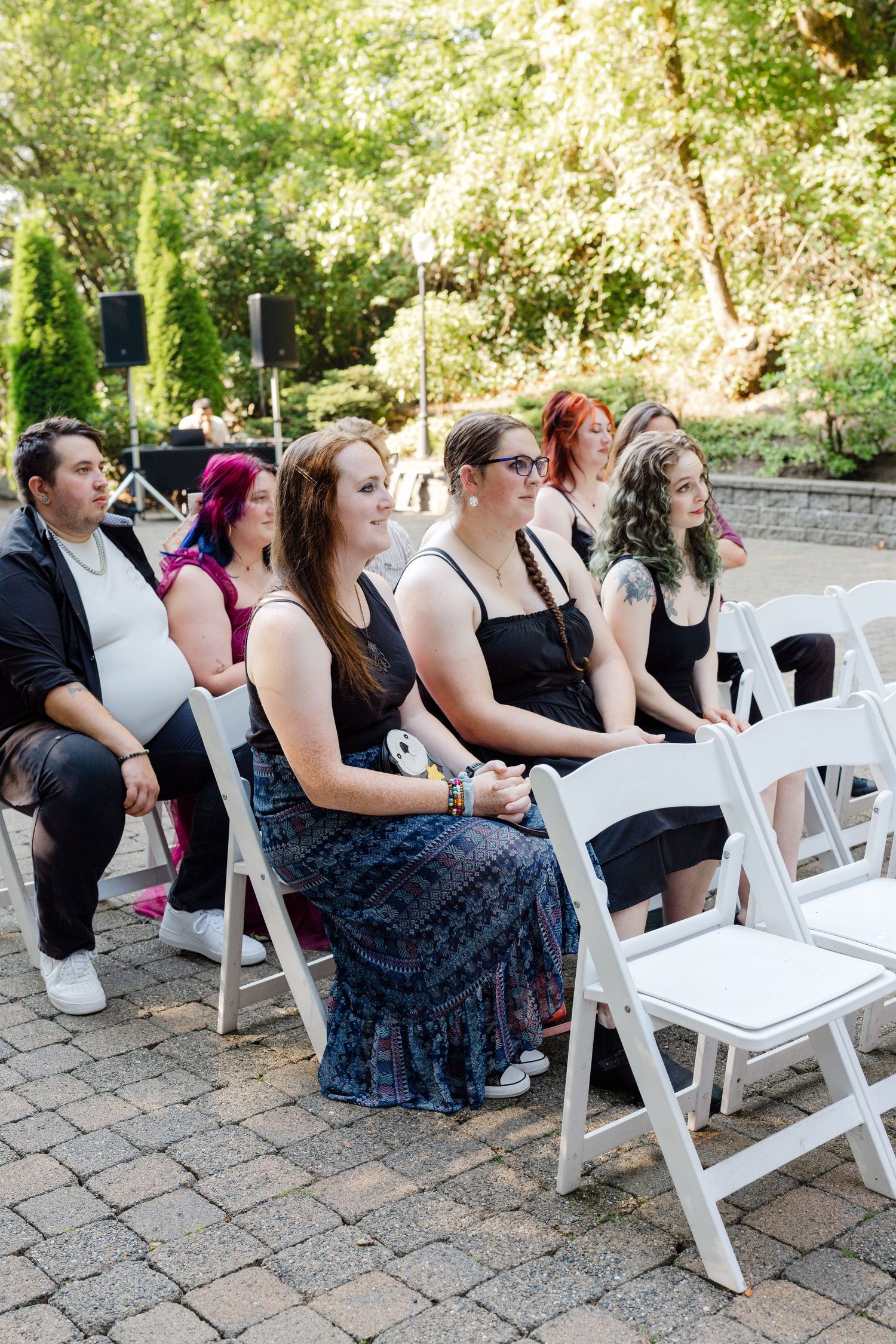 Guests seated in white folding chairs at an outdoor wedding ceremony surrounded by lush green trees.