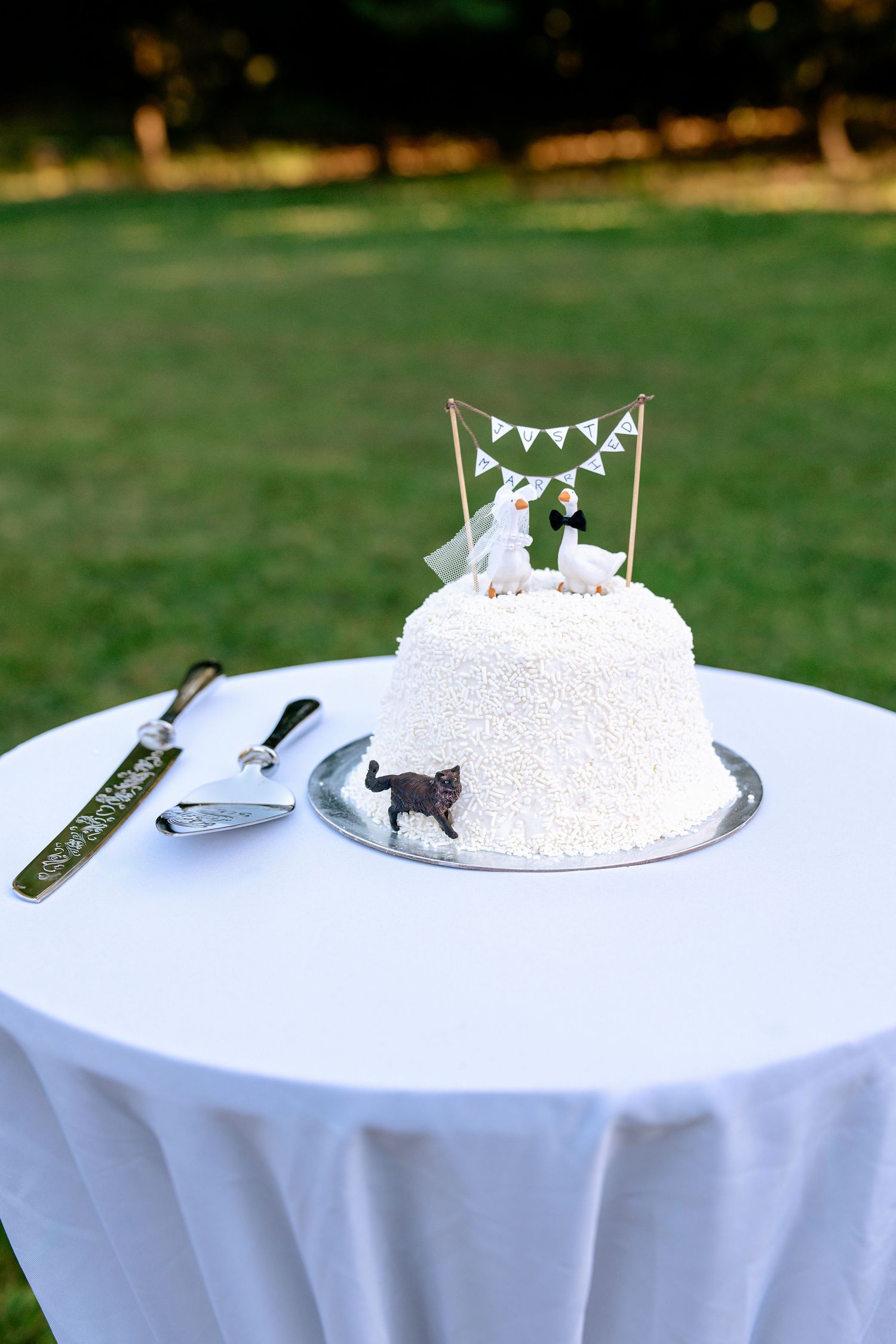 White coconut wedding cake with bride and groom topper, bunting, and black cat figurine on table.