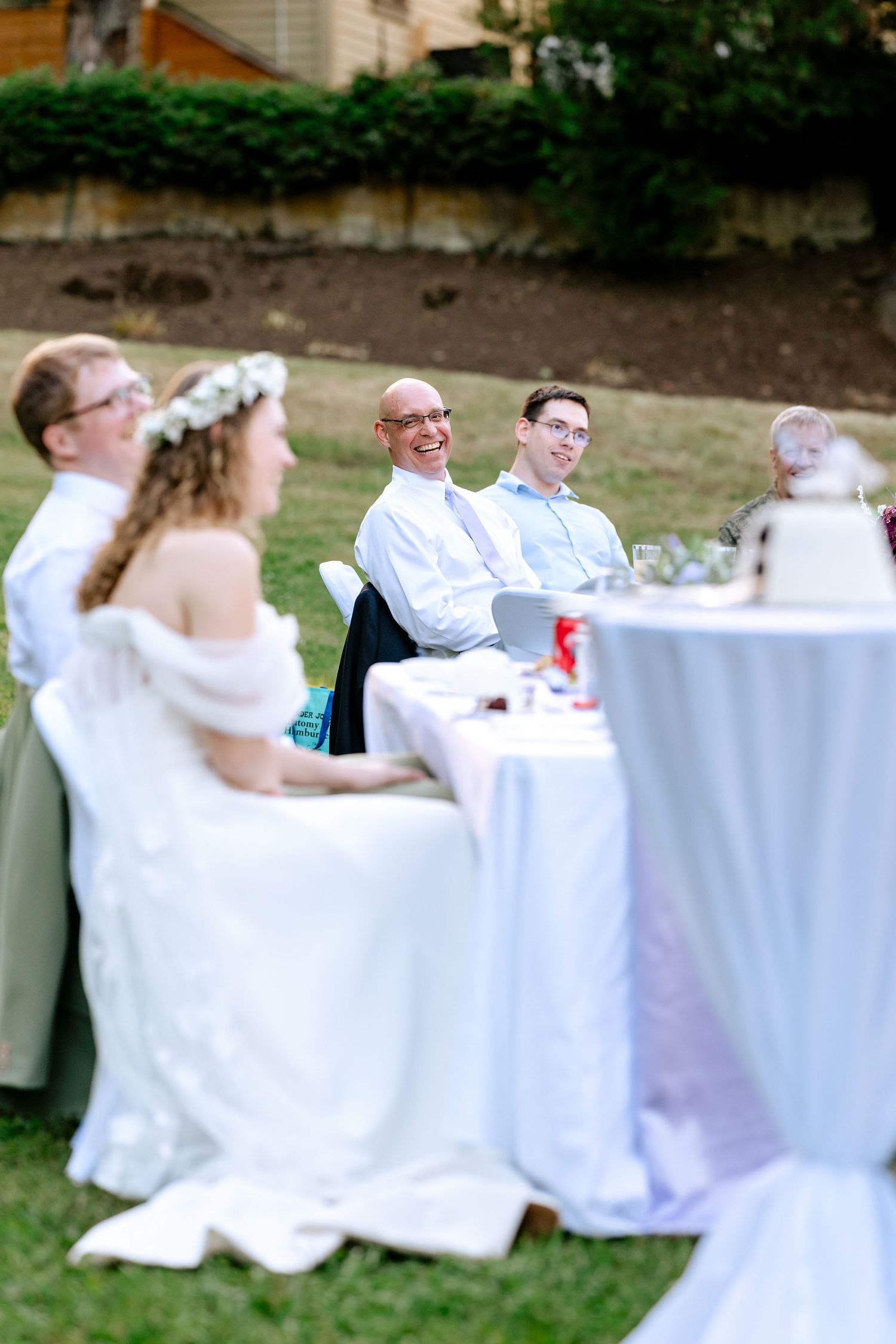 Bride and groom laugh with guests at an outdoor wedding reception table near a white cake stand.