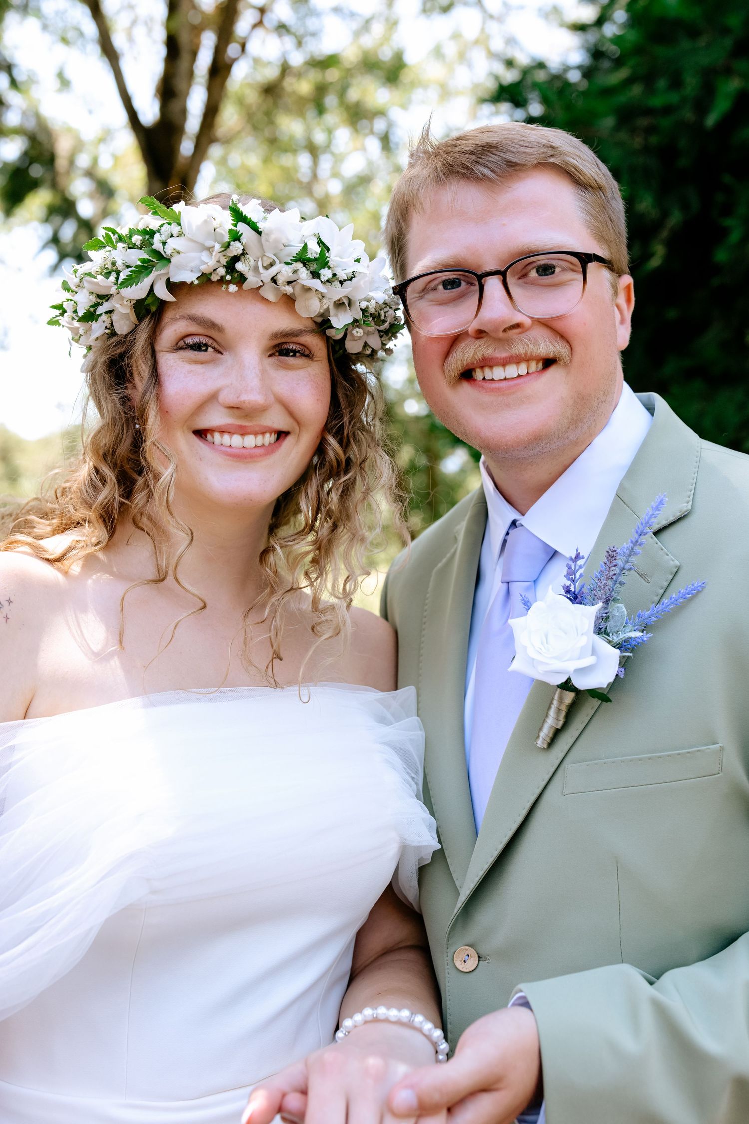 Smiling bride with floral crown and groom in sage suit pose together outdoors on their wedding day.