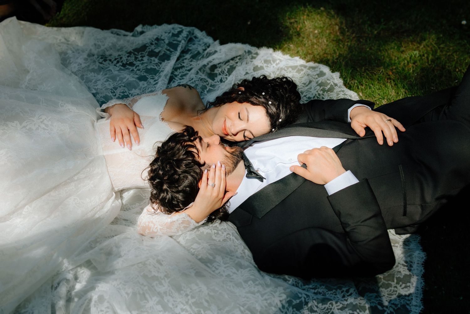 Bride and groom lying on grass in wedding attire, sharing a romantic moment with bride resting her head on groom's chest.