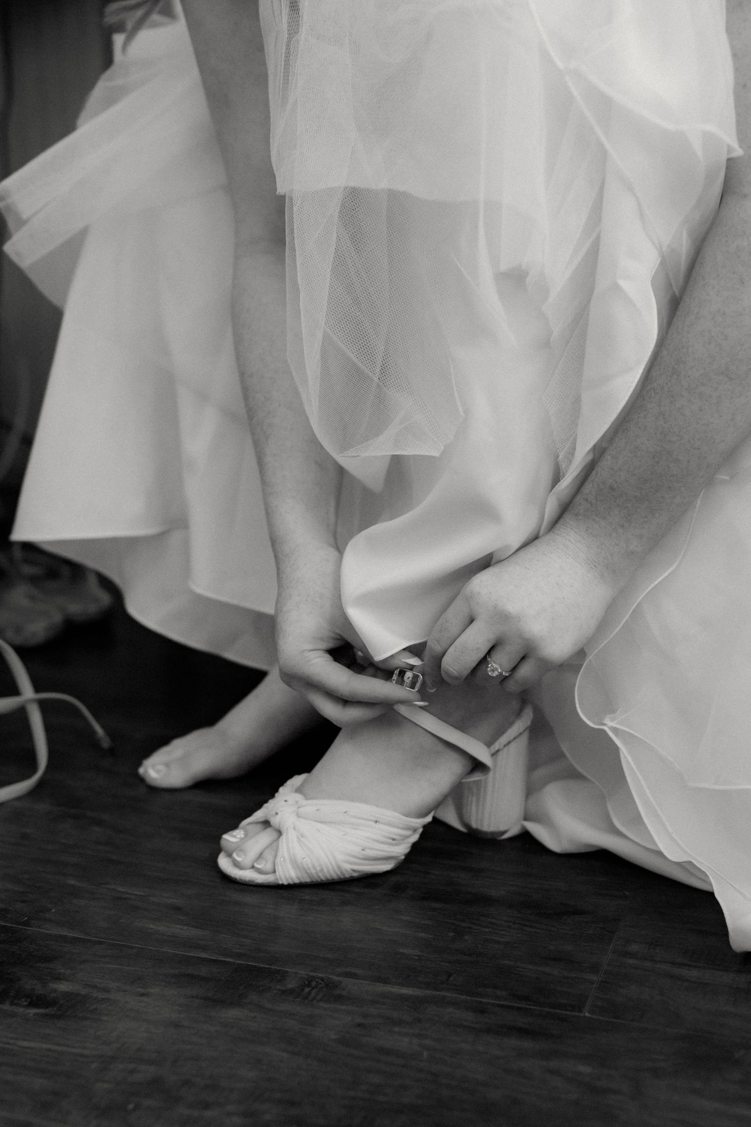 Bride fastening a white heeled wedding shoe in a black and white photo.