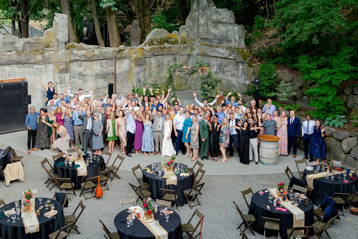 Large group of wedding guests pose together waving in decorated outdoor stone courtyard reception venue.