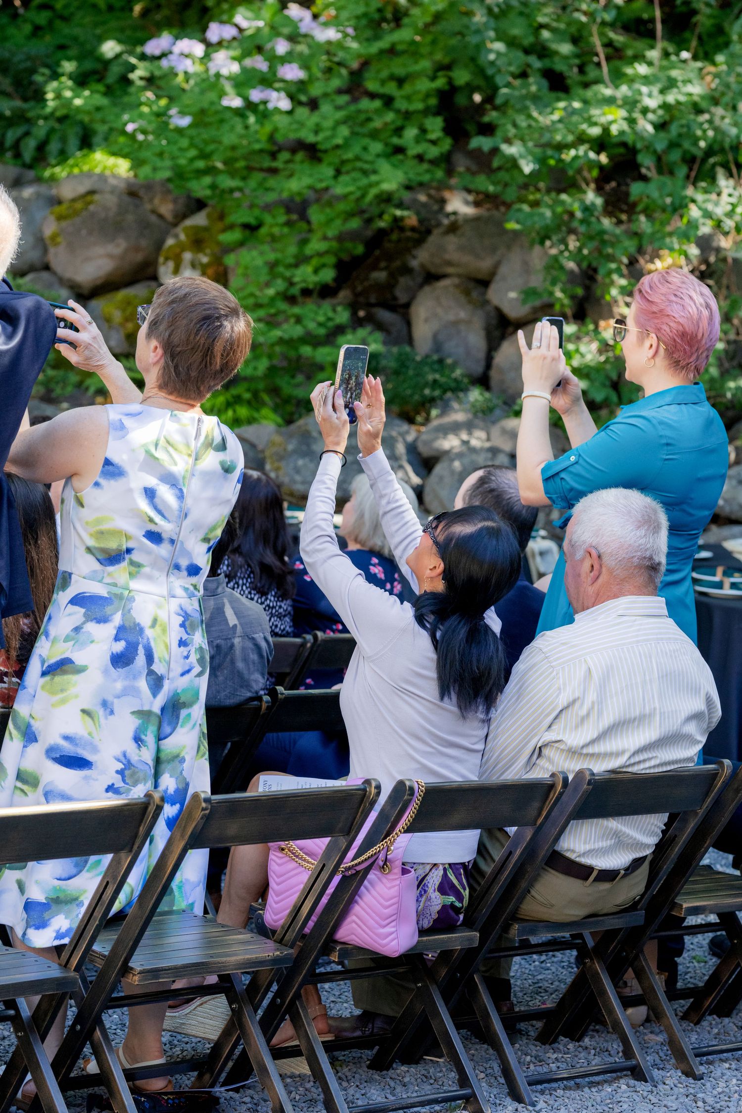 Wedding guests seated outdoors raise phones to capture ceremony moments in a lush garden setting.