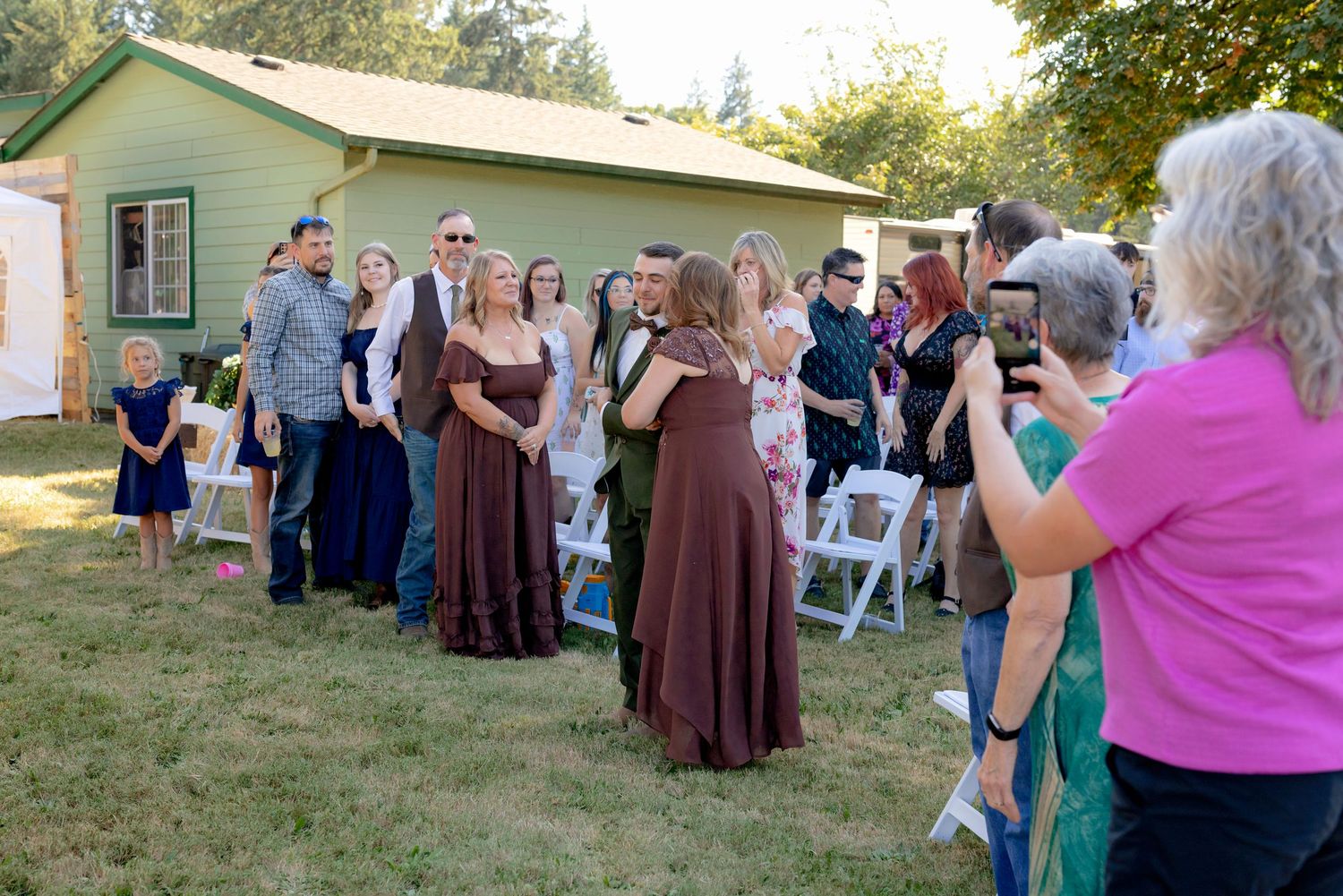 Wedding party in brown and green attire posing for photos outside green building with white chairs nearby.