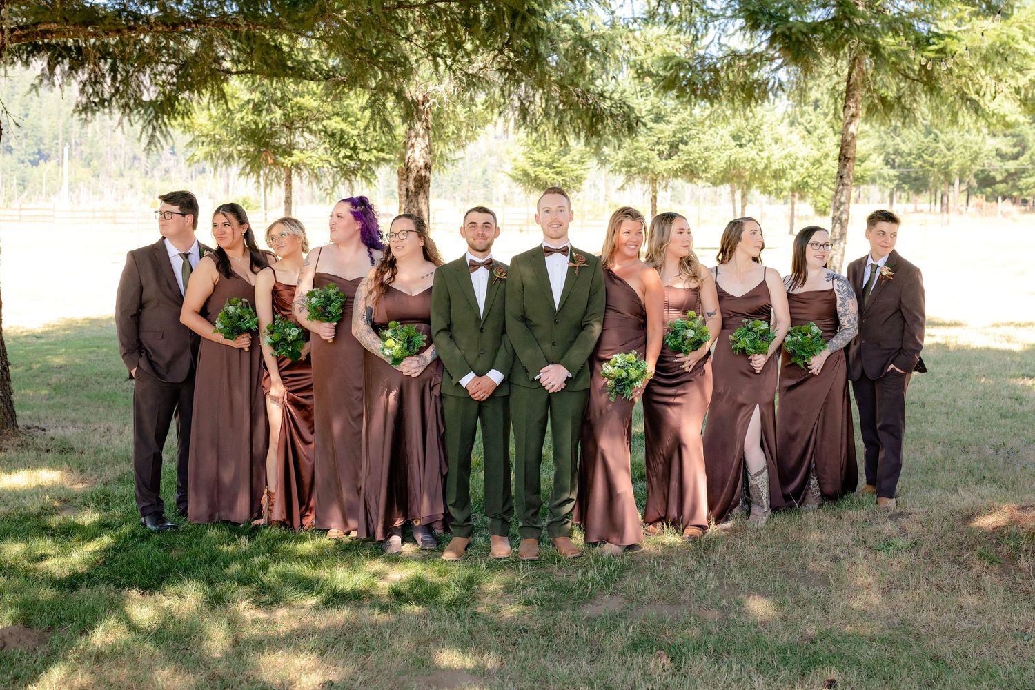 Groom stands with large wedding party in matching brown dresses and suits under pine trees outdoors.
