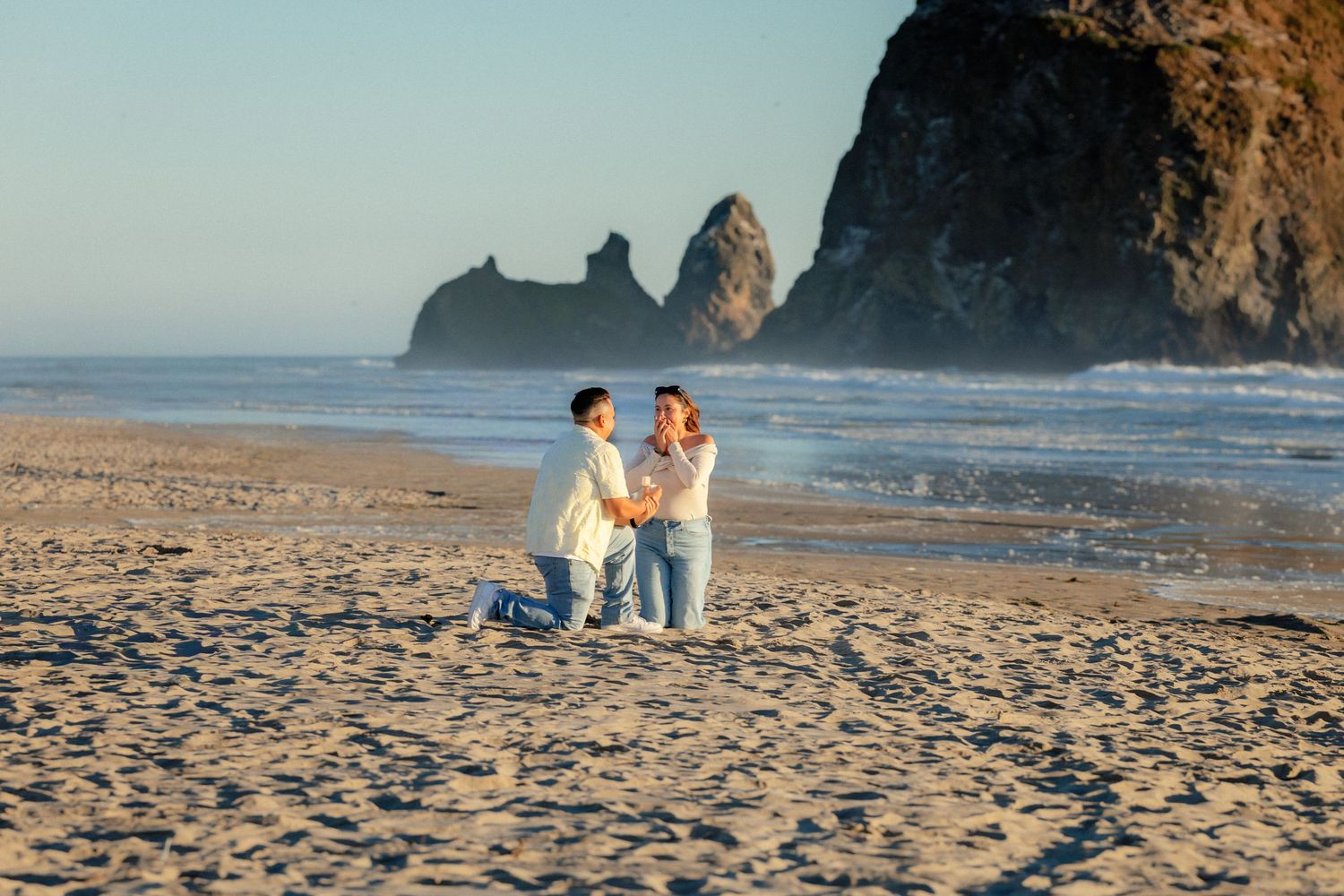 Man proposing to woman on a sandy beach at sunset with dramatic sea stacks in the background.