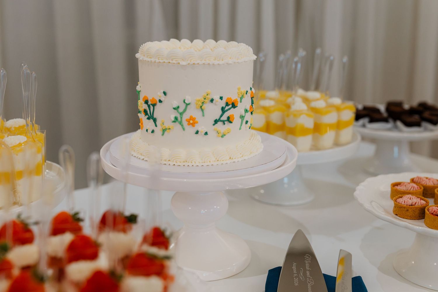 Elegant white wedding cake with floral embroidery design displayed on a dessert table with treats.