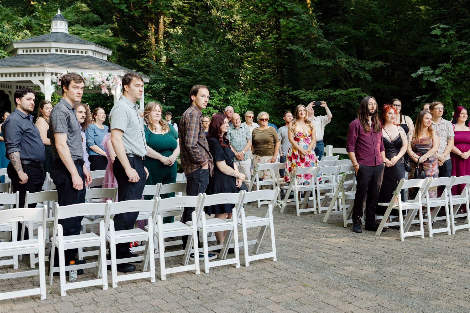 Wedding guests stand and watch outdoor ceremony near white gazebo surrounded by tall green trees.