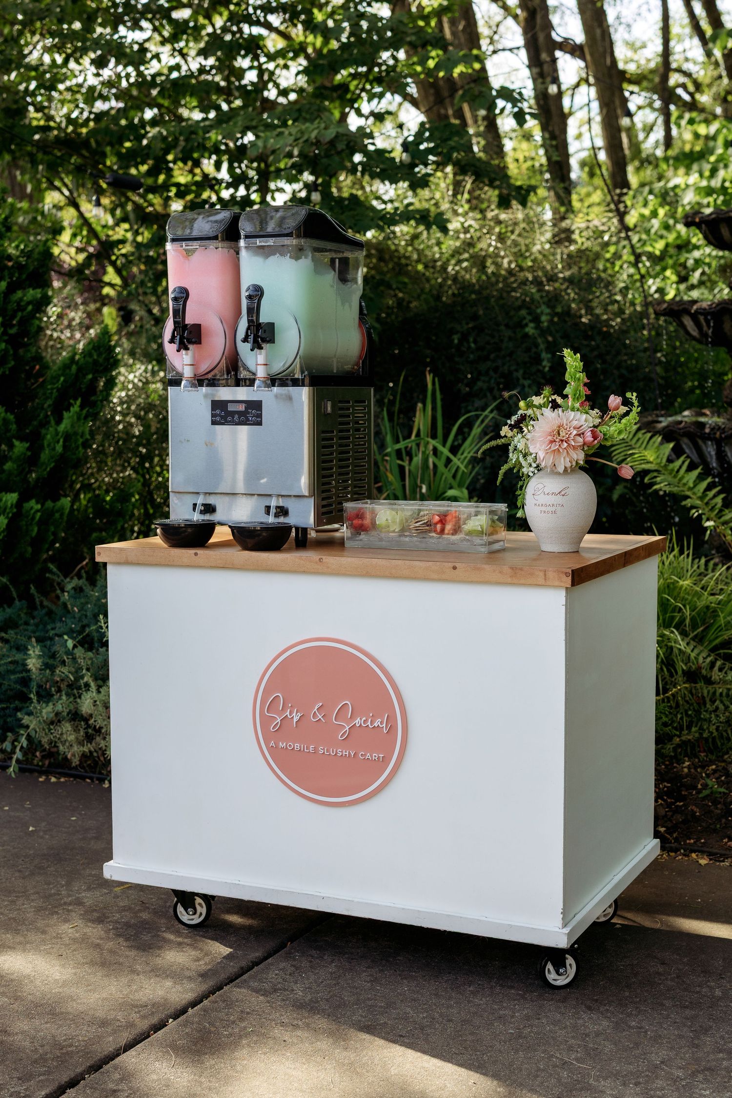 Mobile white bar cart with wooden countertop and branded signage in an outdoor garden setting.