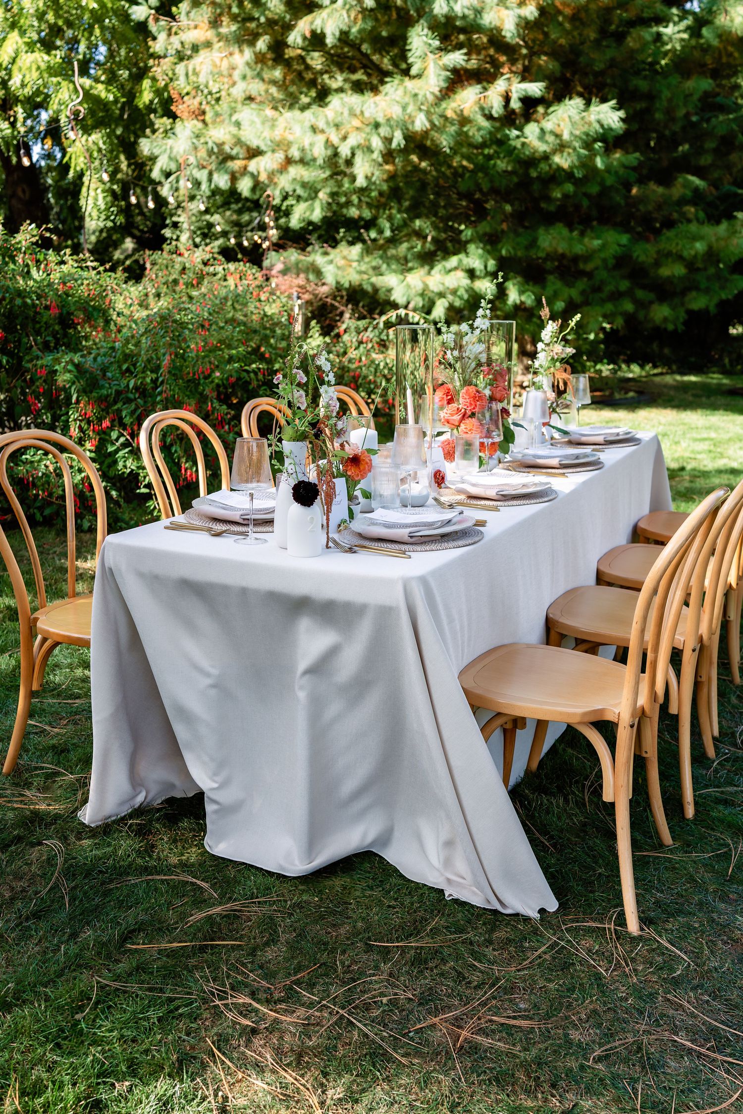Elegant outdoor wedding reception tables with white linens and wooden chairs set beneath trees.