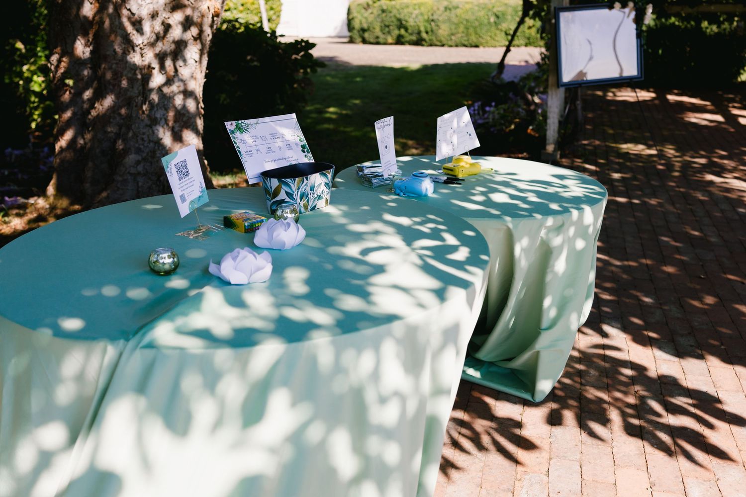 Wedding reception tables decorated in soft mint green linens with place settings under dappled sunlight.