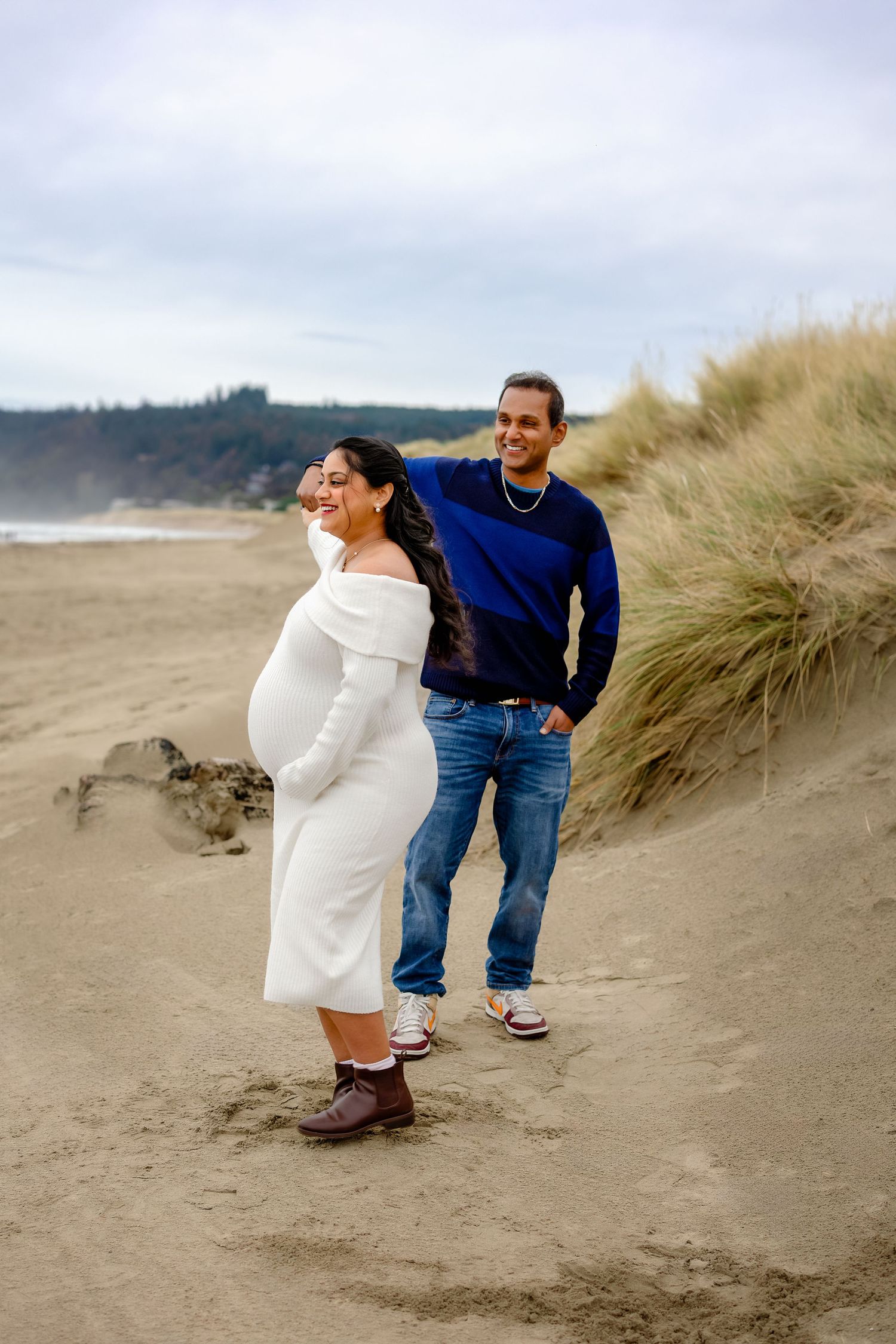 A couple shares a playful moment among sand dunes at the beach on an overcast day.