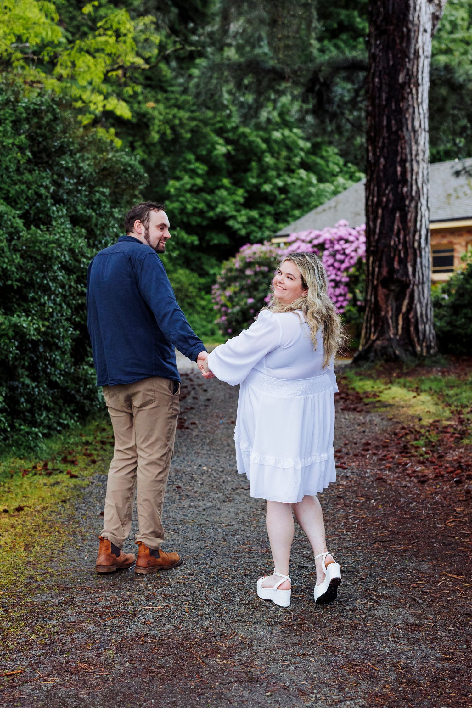 A couple walks hand in hand down a forest path with blooming flowers and greenery.