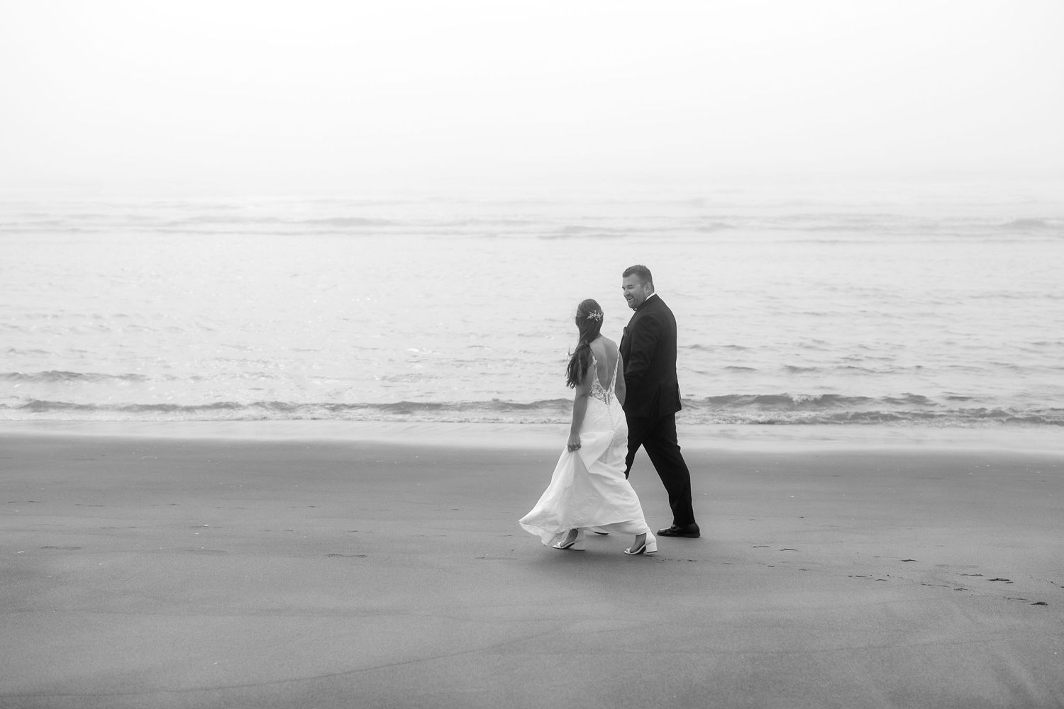 Black and white photo of newlyweds walking together along a misty beach shoreline.
