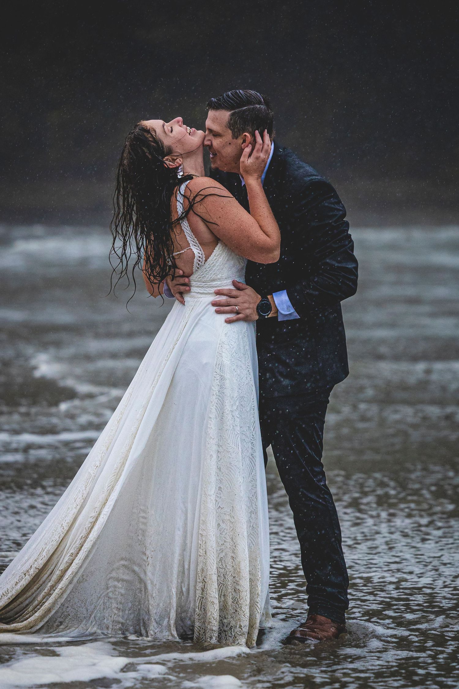 Romantic wedding couple shares passionate kiss in shallow ocean water with wet wedding dress trailing in waves.