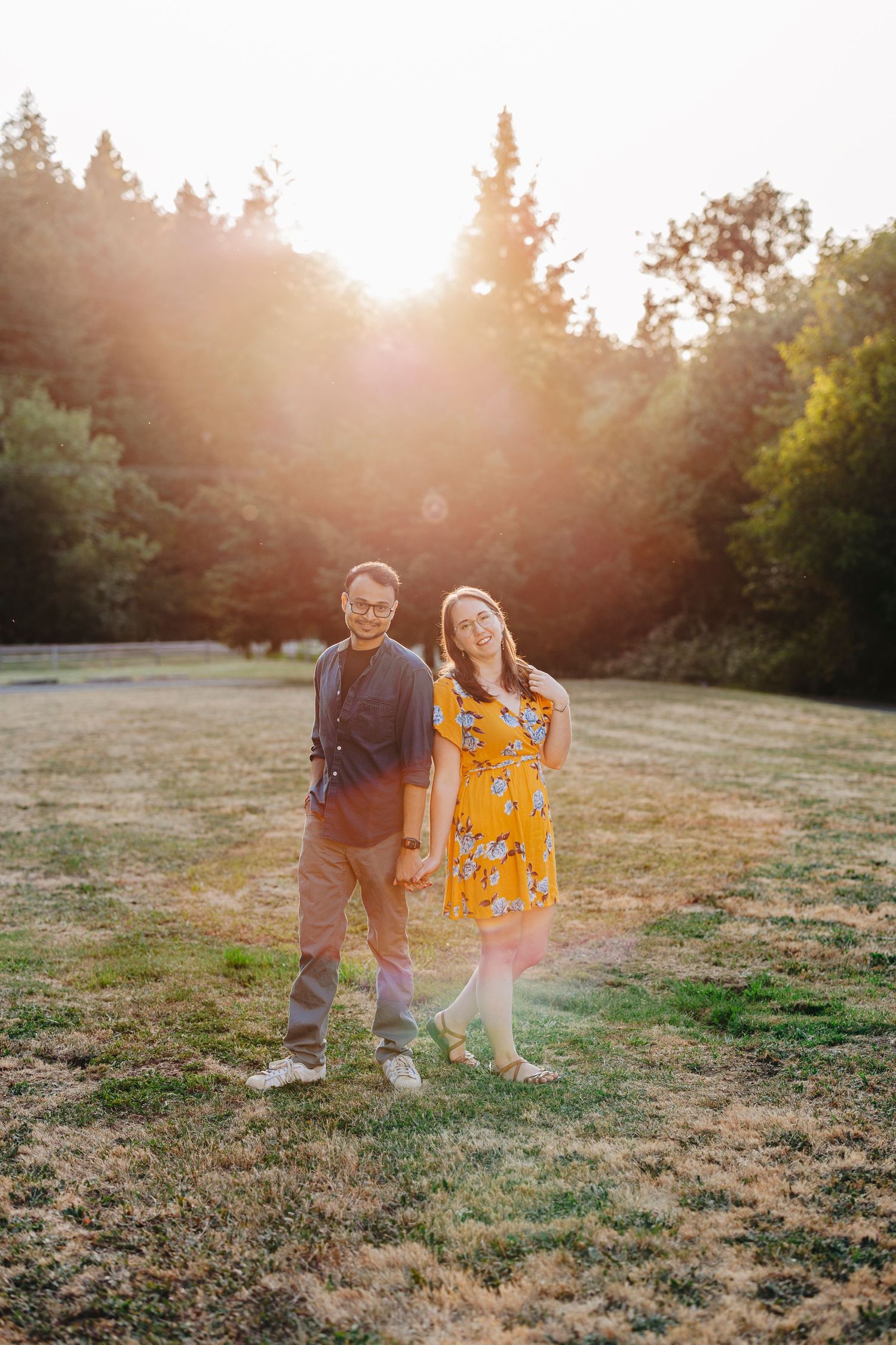 Couple walking through golden sunset light in an open field with evergreen trees in background.