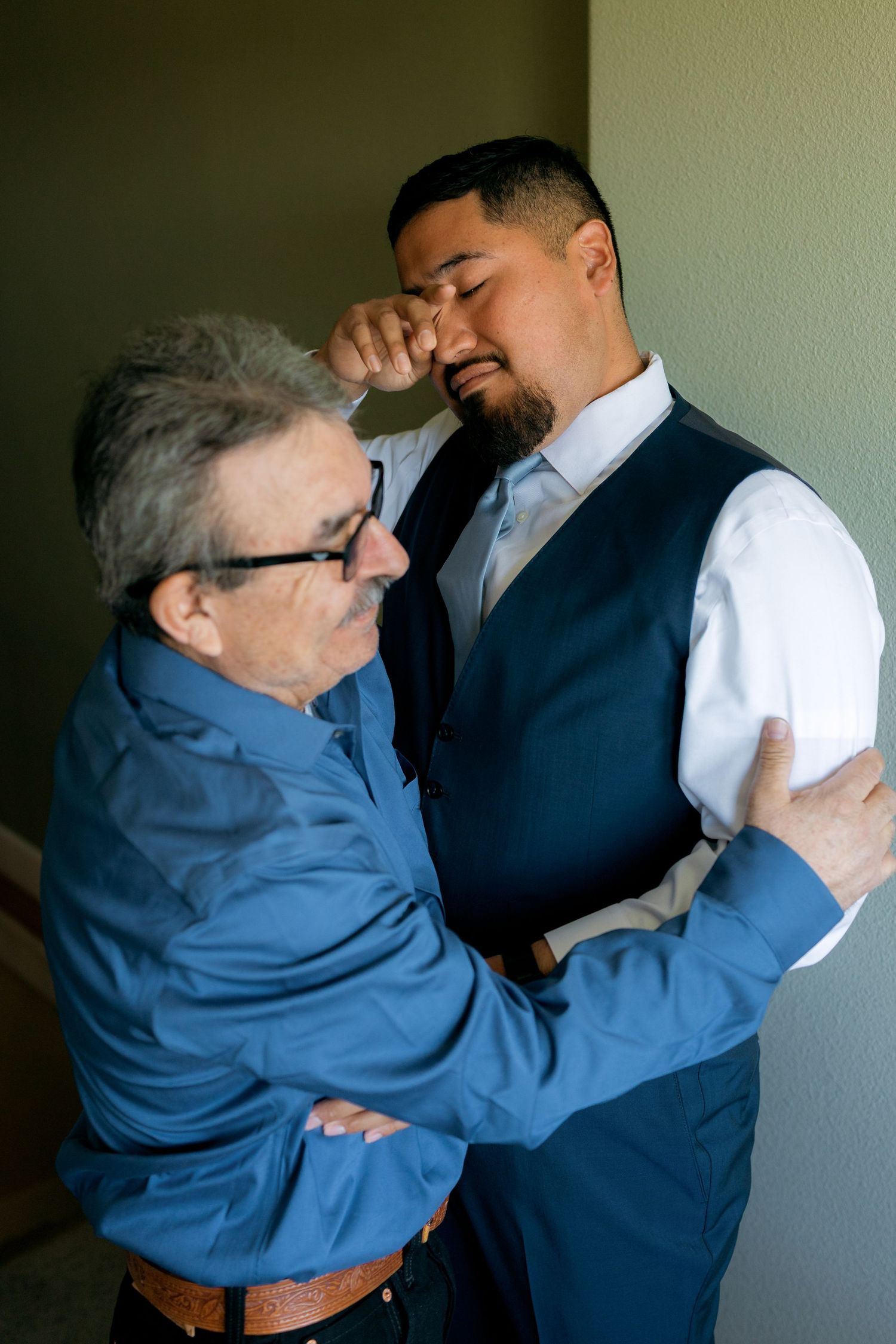 Emotional groom wipes tears as older man embraces him during wedding day preparations indoors.