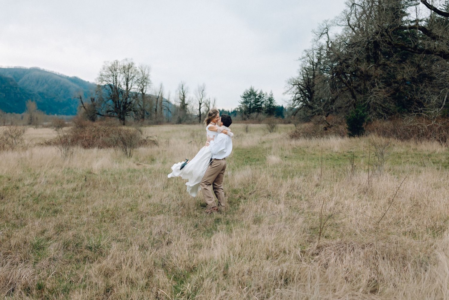 Panoramic wedding scene with couple in an open field against mountain backdrop.