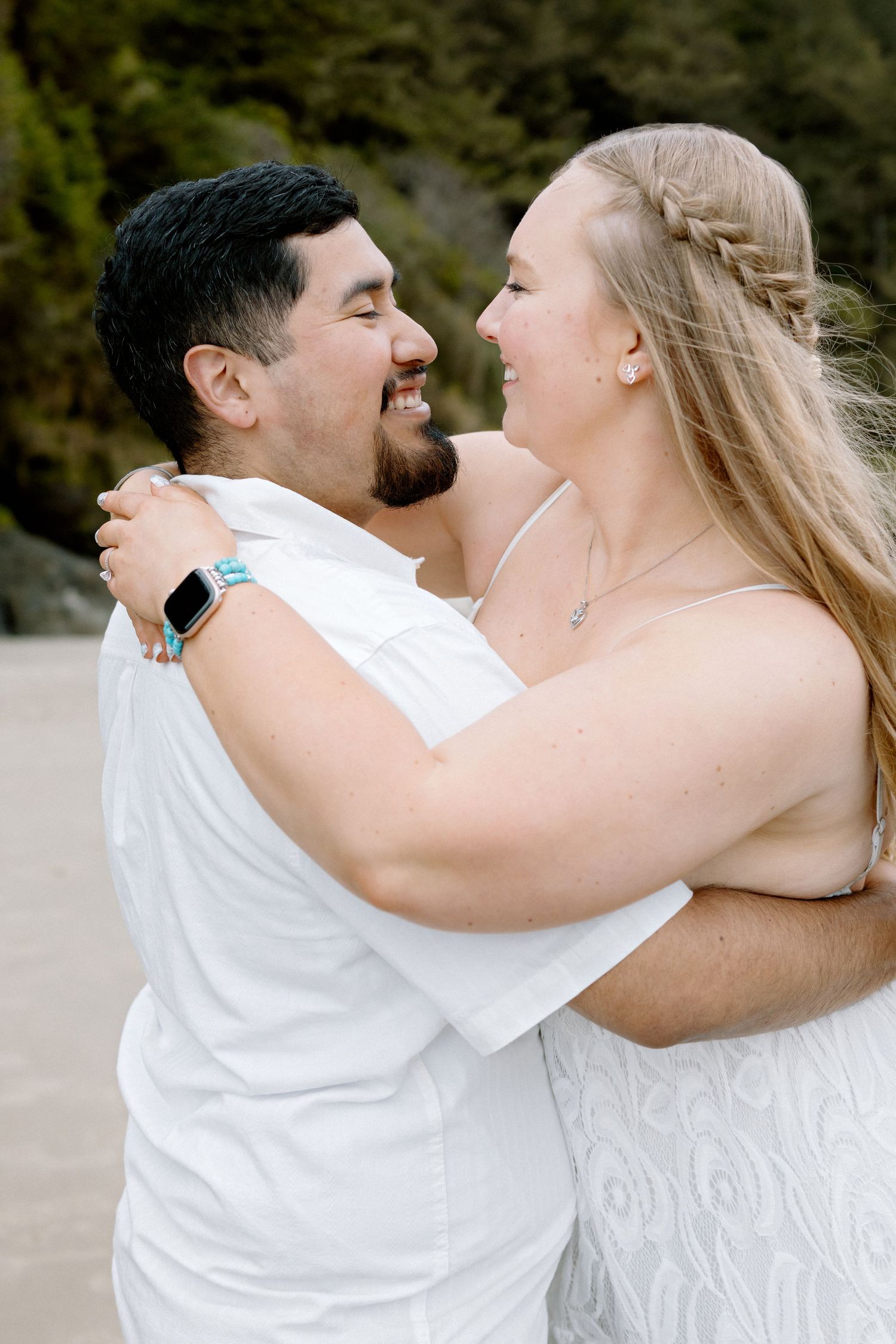 Couple in white outfits embracing and smiling at each other on a beach with trees in background.