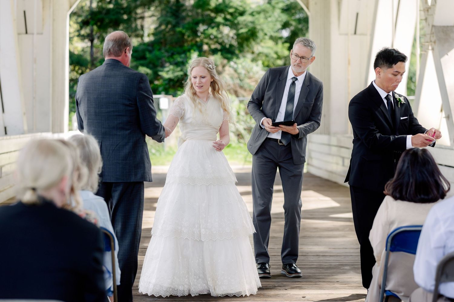 A bride in a white gown walks down the aisle at an outdoor wedding ceremony under a white pavilion.