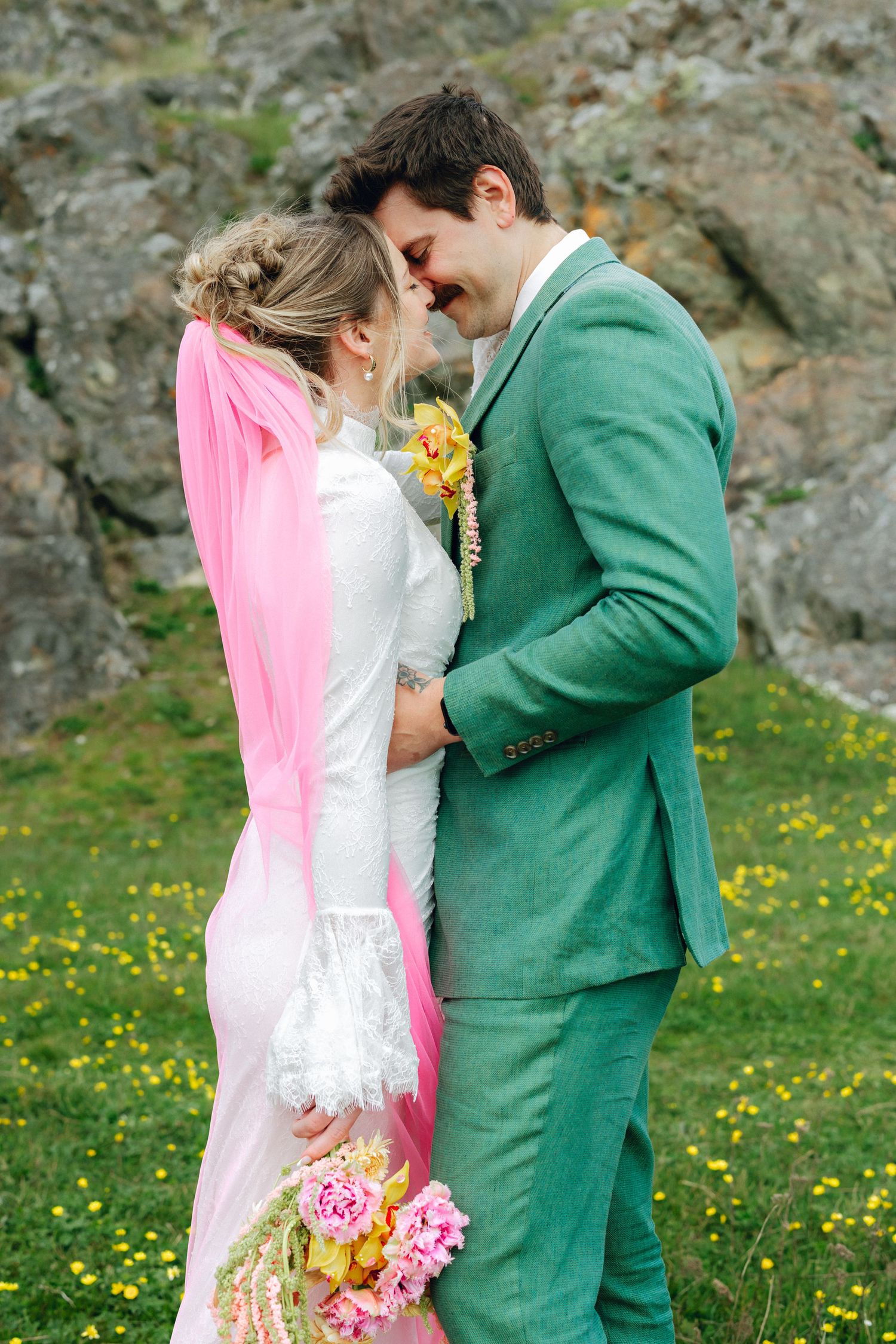 Smiling bride and groom in green suit touching foreheads romantically outdoors with yellow wildflowers and rocky backdrop.