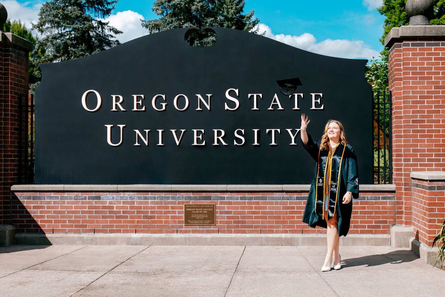 Graduate in cap and gown posing in front of Oregon State University brick entrance sign on a sunny day.