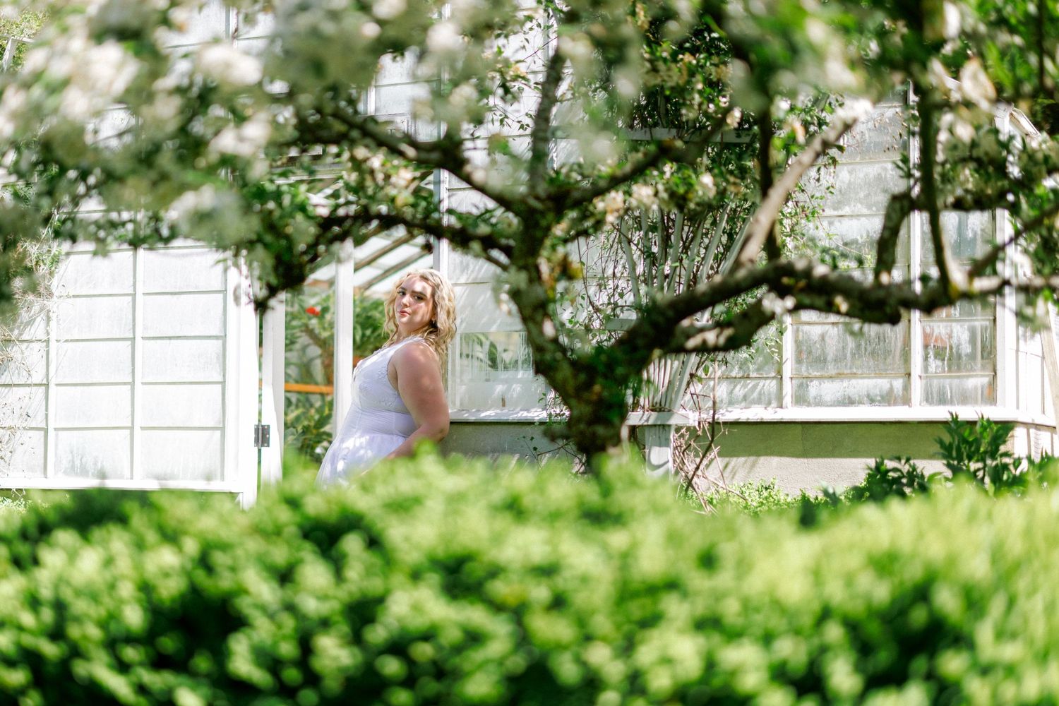 Blonde woman in white dress standing near a greenhouse framed by blooming white tree branches.