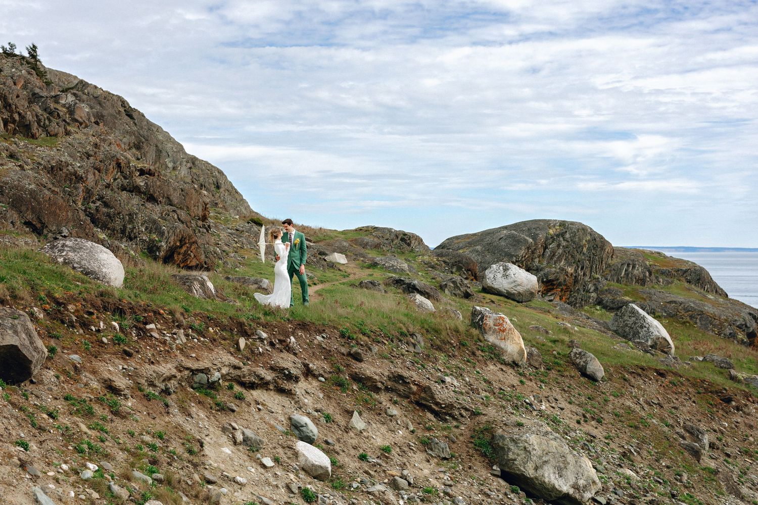 Couple in wedding attire standing on rocky coastal hillside with ocean views and dramatic boulder landscape.