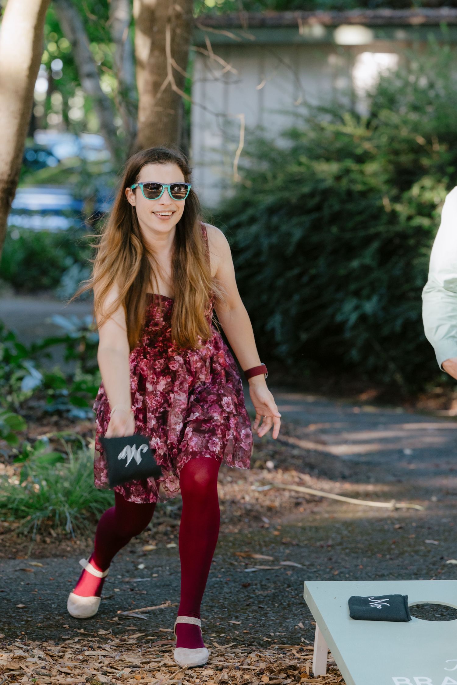 Young woman in floral dress and red tights plays cornhole at an outdoor wedding reception.