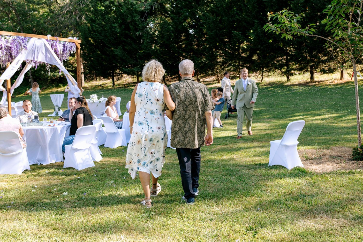 Elderly couple walks hand in hand across a sunny lawn toward a decorated outdoor wedding reception area.