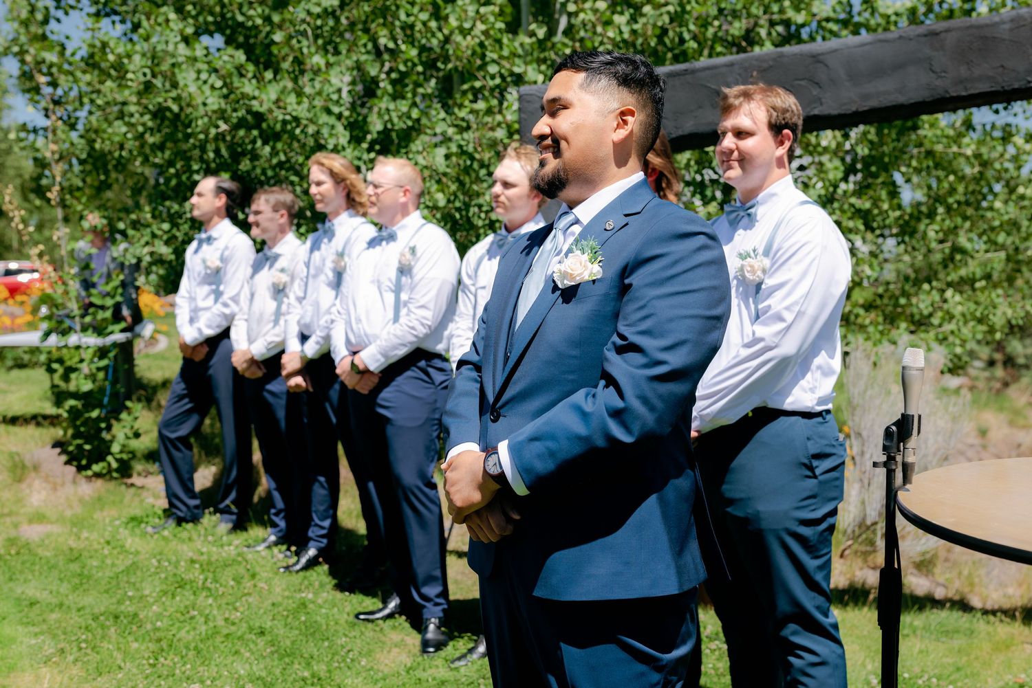 Groom in navy suit with boutonniere stands with groomsmen in white shirts at outdoor wedding ceremony.