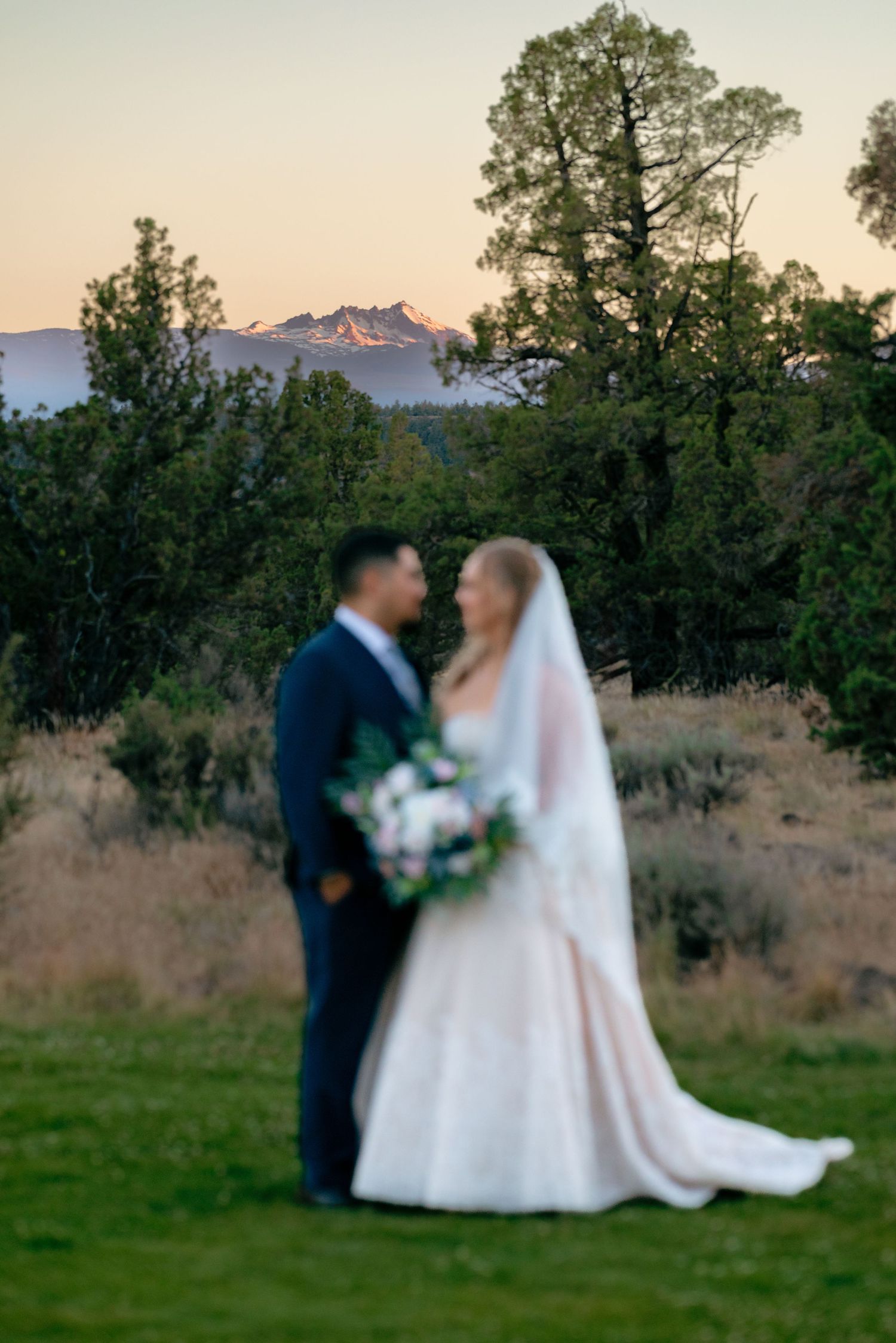 Blurred bride and groom share romantic moment at sunset with snow-capped mountain backdrop.