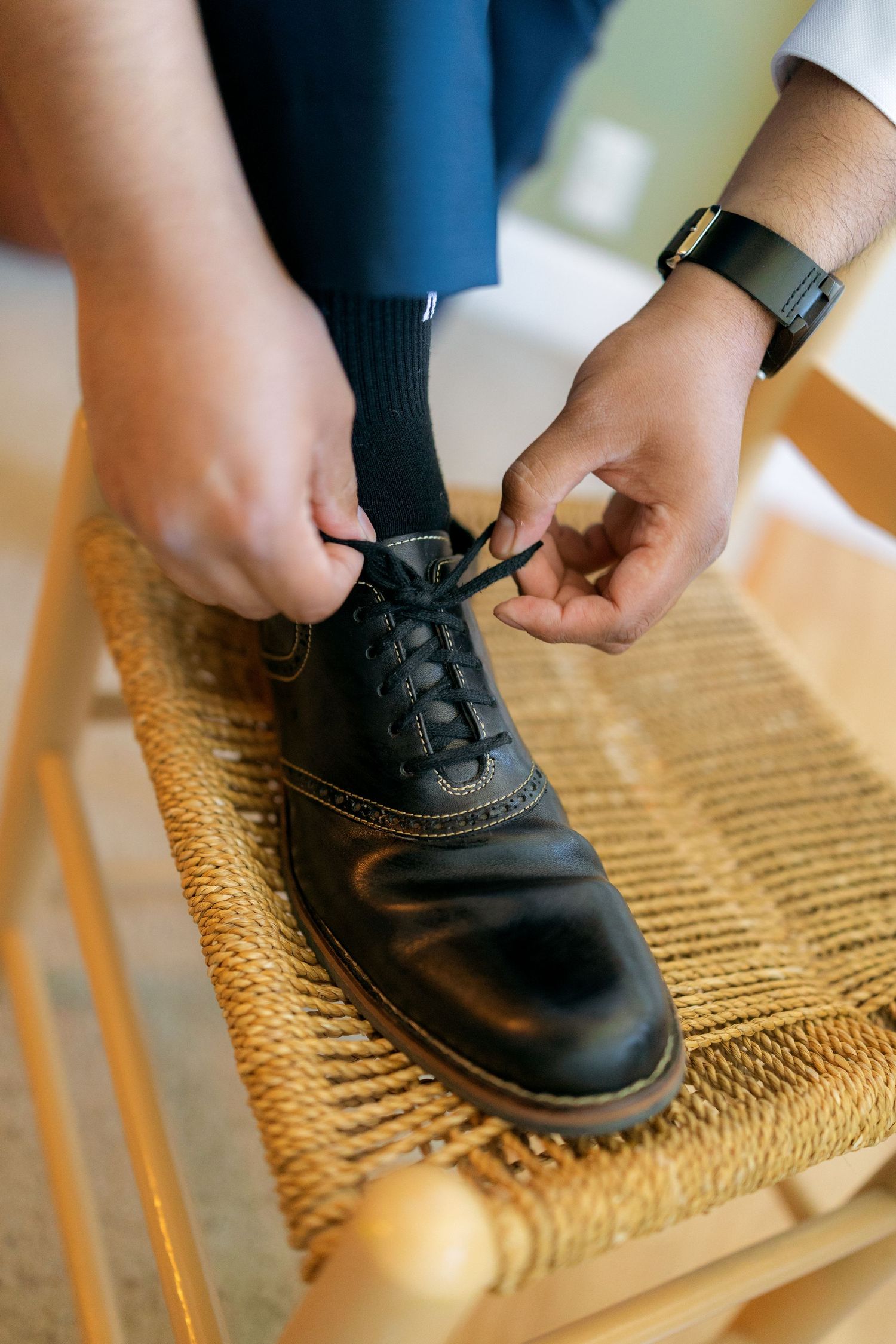 Groom tying black dress shoe laces on a woven chair, wearing navy blue trousers and a smartwatch.