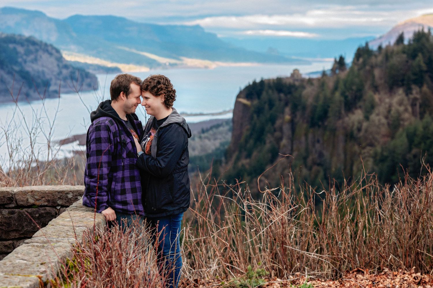 A couple shares an intimate moment at a scenic overlook with mountains and a lake in the background.