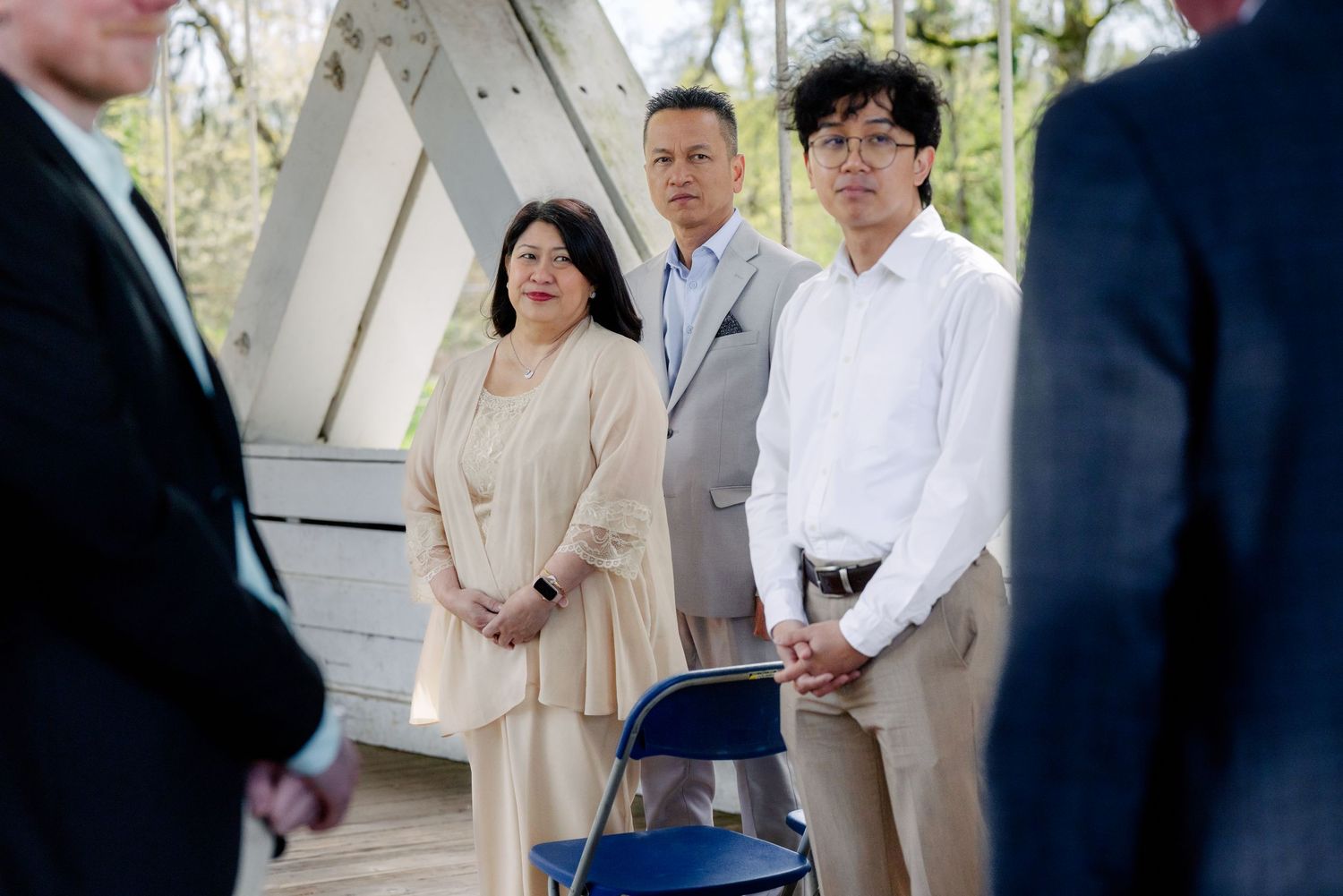 A family in formal attire stands attentively outdoors at a wedding ceremony near a white wooden structure.