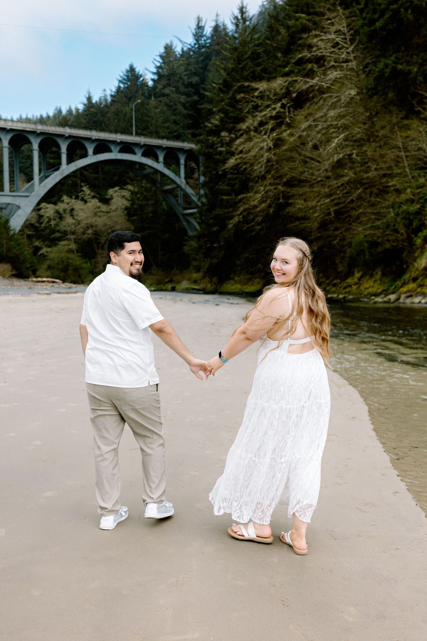 Smiling couple holding hands on beach with scenic arched bridge and evergreen forest backdrop.