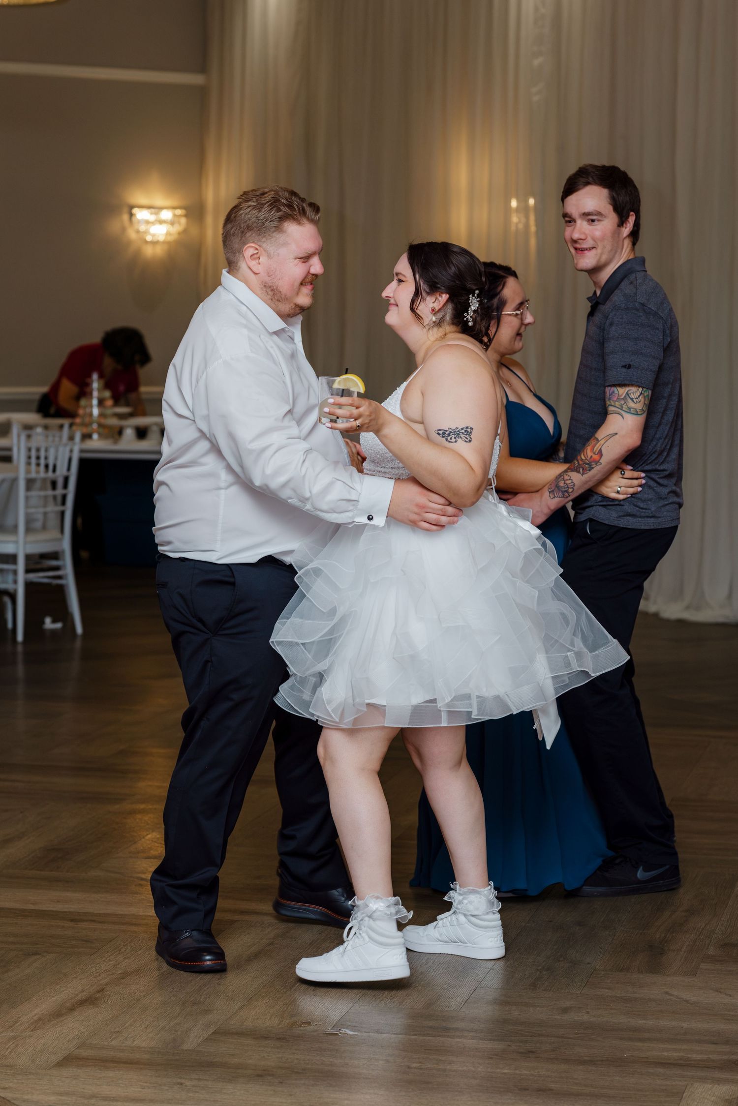 Bride in short white dress dances joyfully with guests at indoor wedding reception on wooden dance floor.