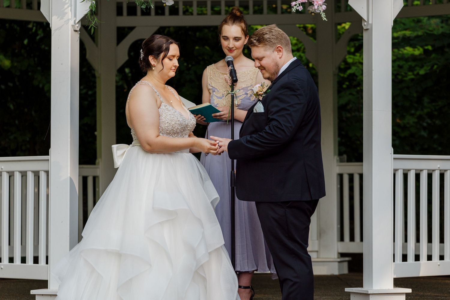 Bride and groom exchange vows under a white gazebo, officiated by a woman in lavender dress, surrounded by lush greenery.