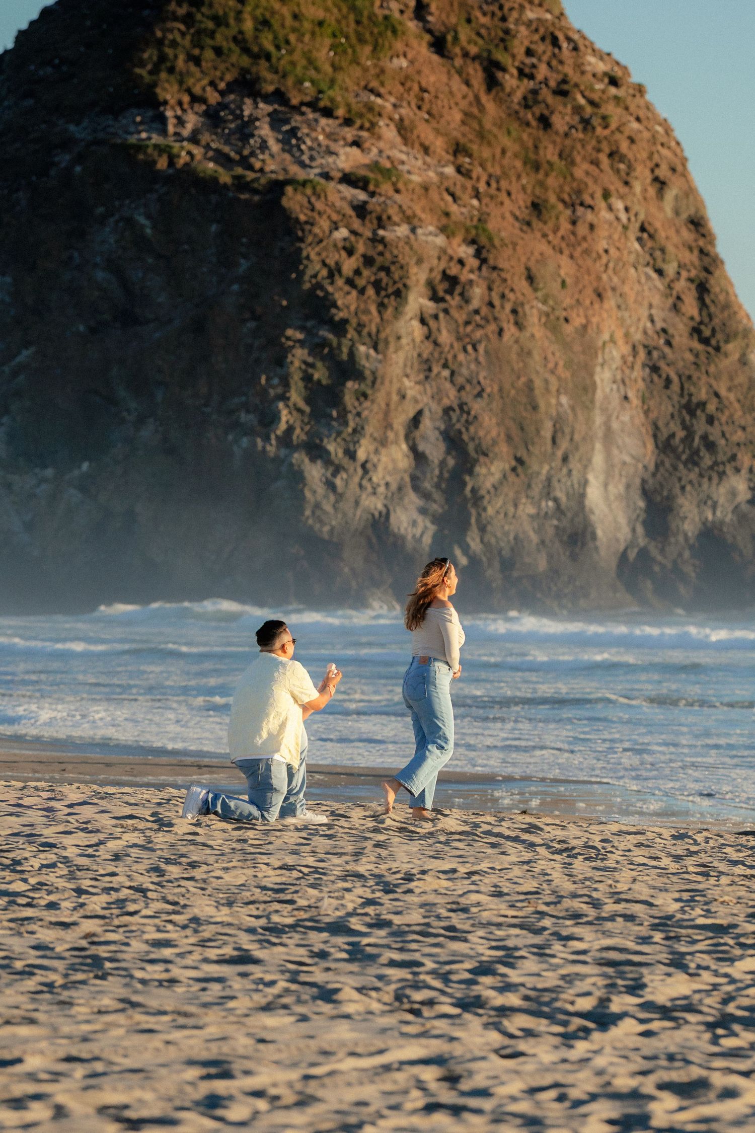 Man proposes on bended knee to woman on sandy beach with dramatic rock formation and ocean waves at sunset.