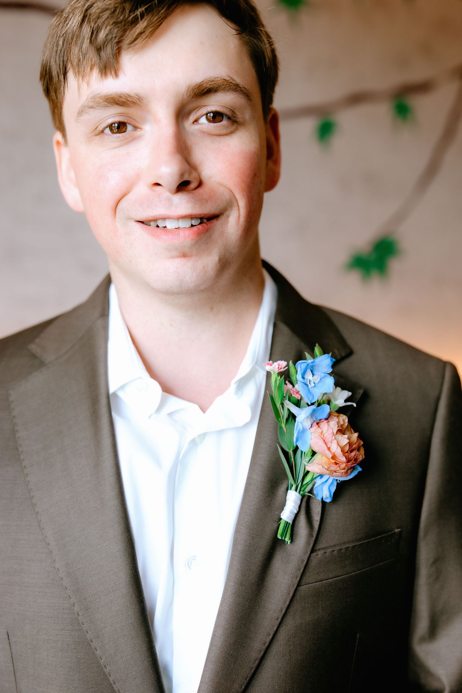 Smiling groom in brown suit with blue and pink floral boutonniere on wedding day.