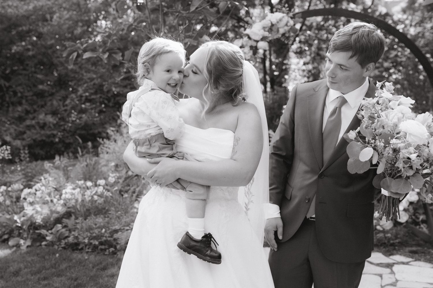 Black and white photo of bride and groom kissing smiling toddler at garden wedding.