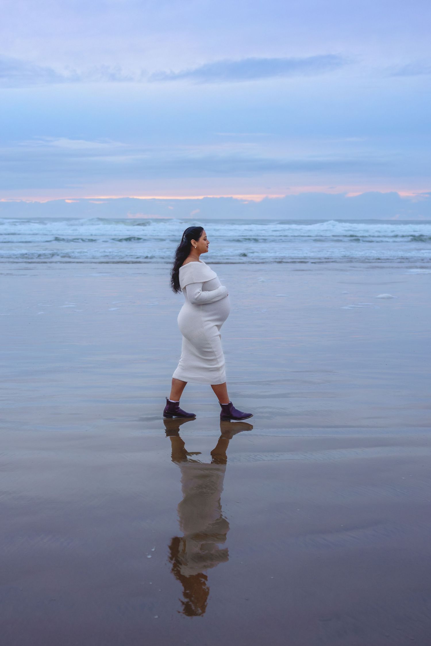 Person in white sweater poses reflectively at the ocean's edge during sunset.