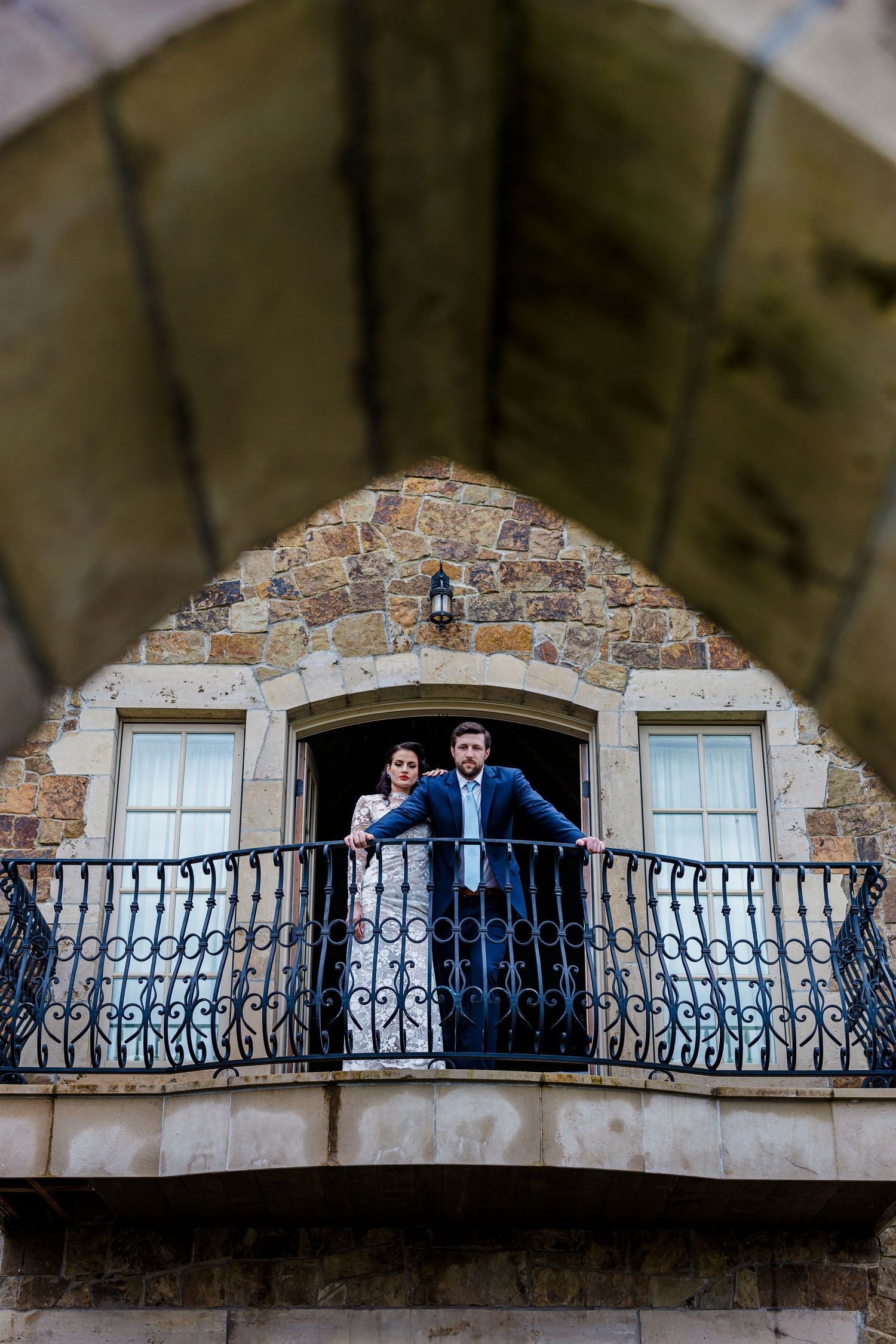 A couple stands together on an ornate stone balcony with wrought iron railings of a rustic building.