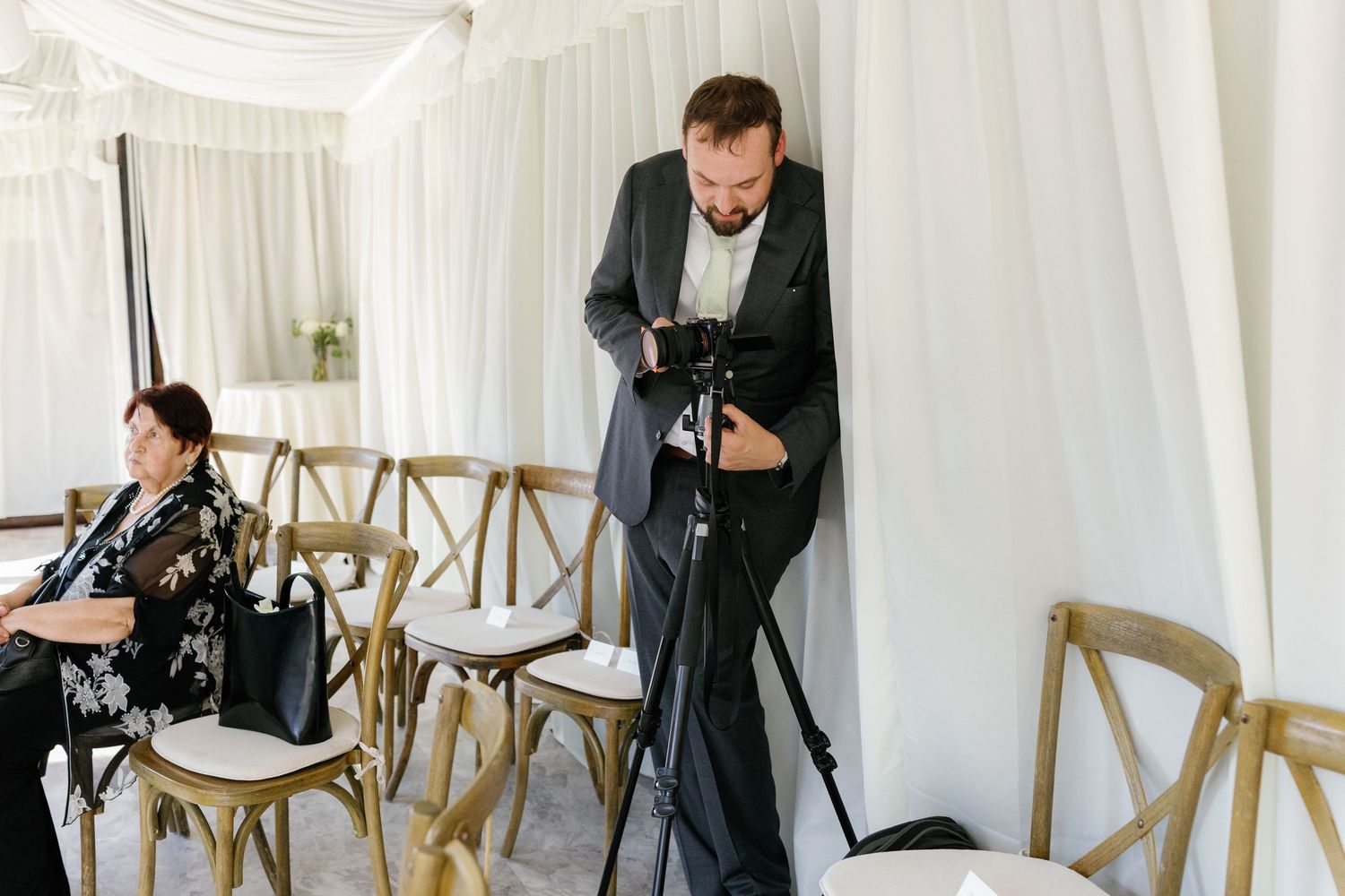 Wedding ceremony setup with white draping and wooden crossback chairs in an indoor venue.