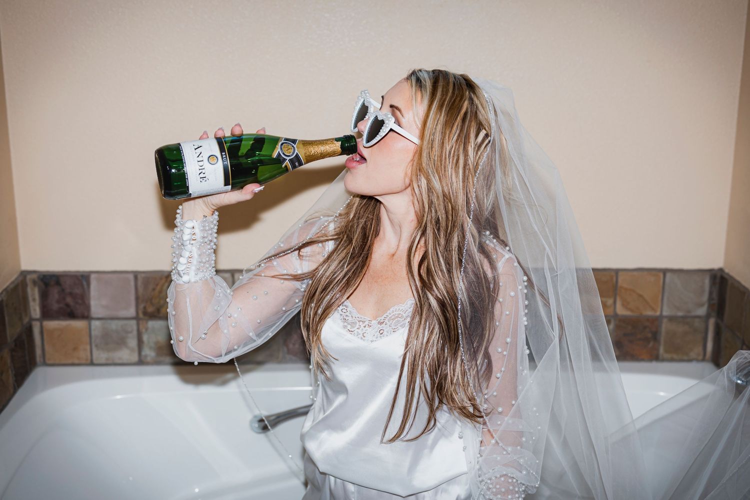 A bride in a white dress and veil sits in a bathtub while drinking from a champagne bottle.