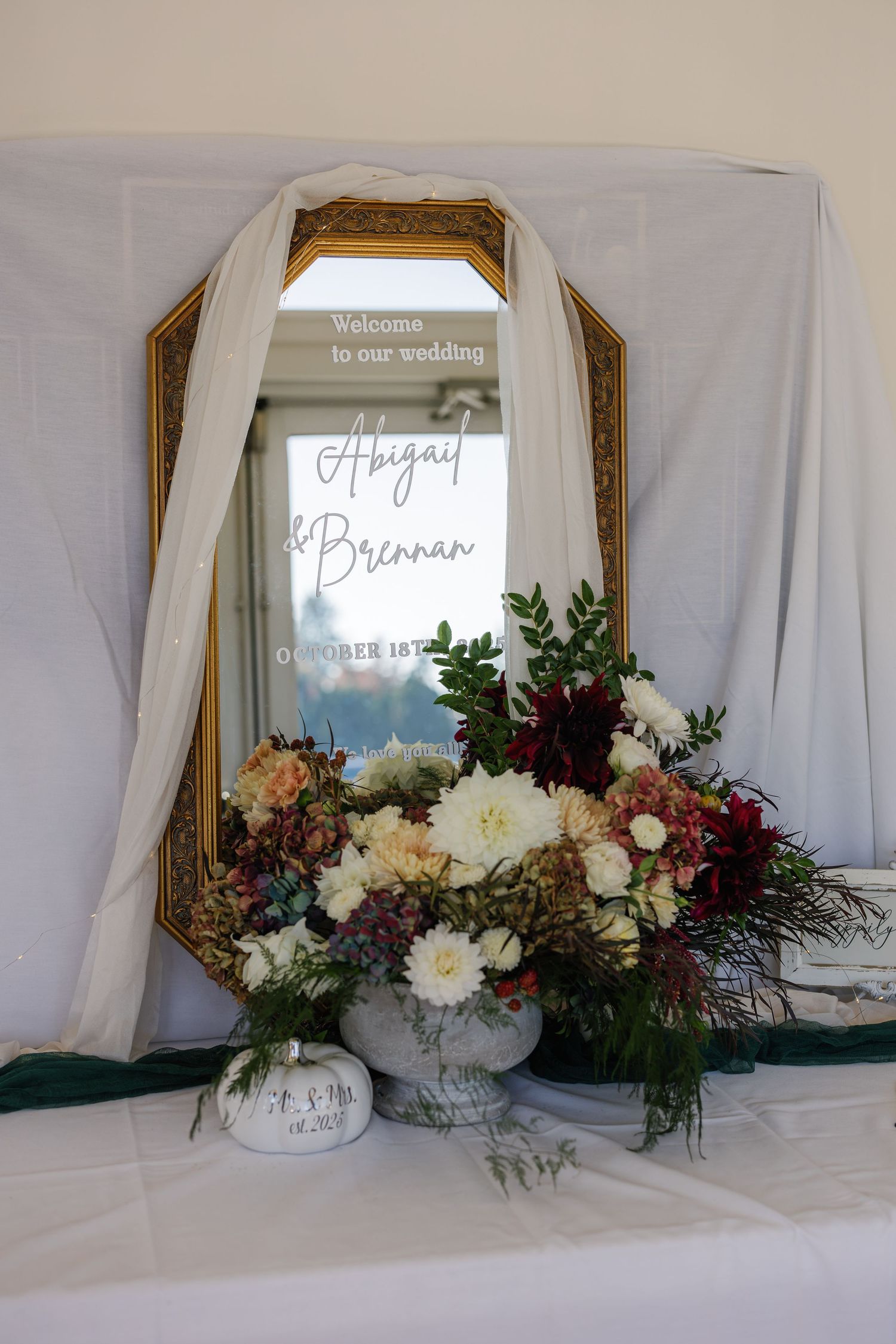 Elegant wedding welcome display with ornate gold mirror and floral arrangements in white and burgundy.